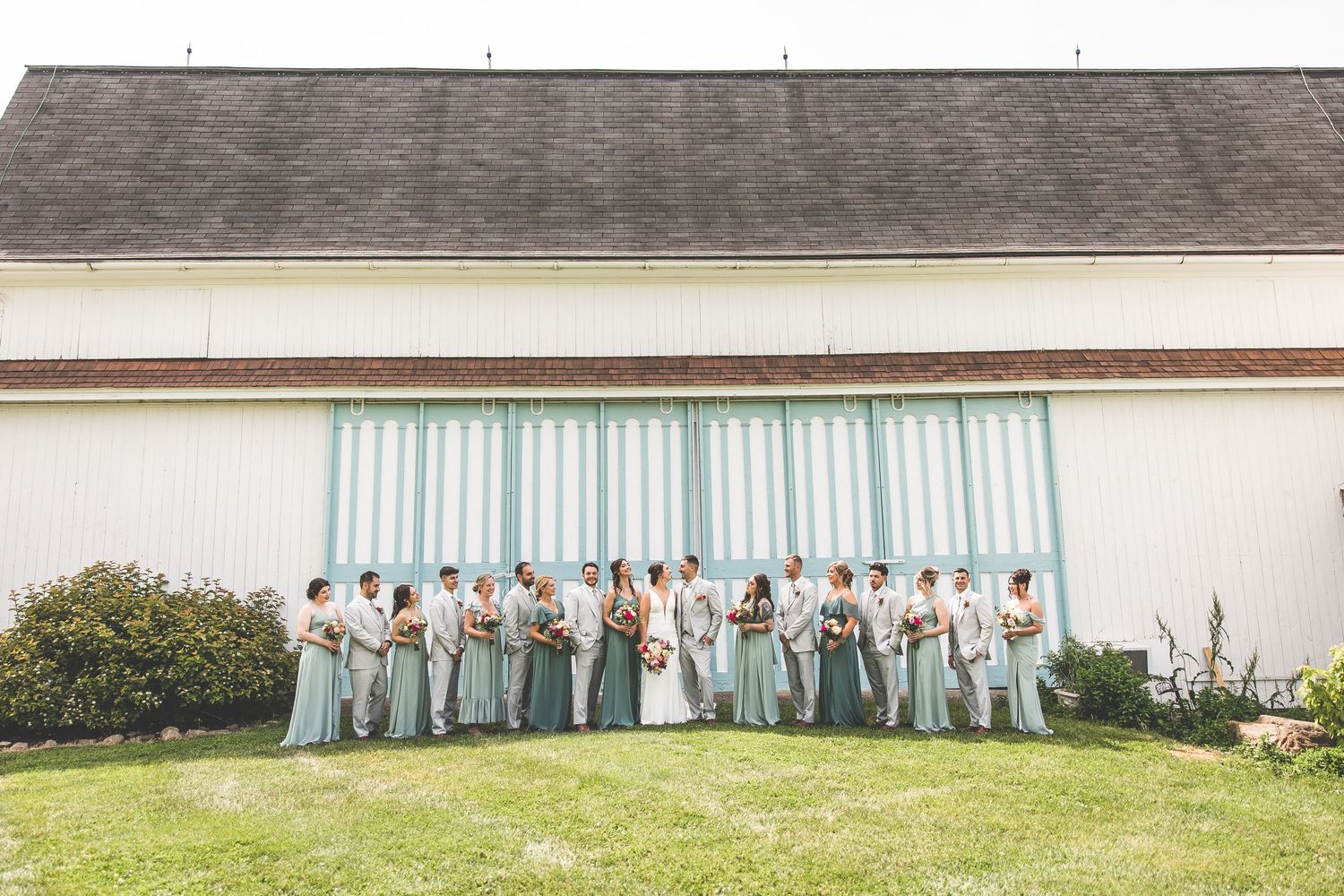 Wedding party lines up against a white building with sage green and gray attire.