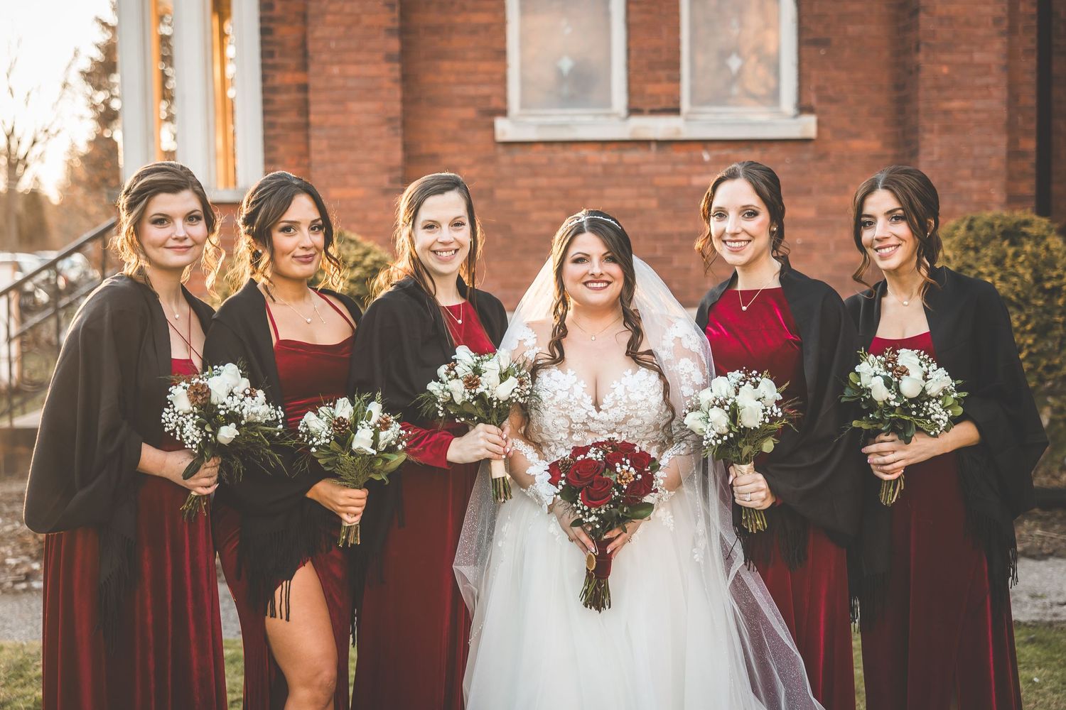 Wedding party in burgundy dresses and black suits pose together outside brick church.