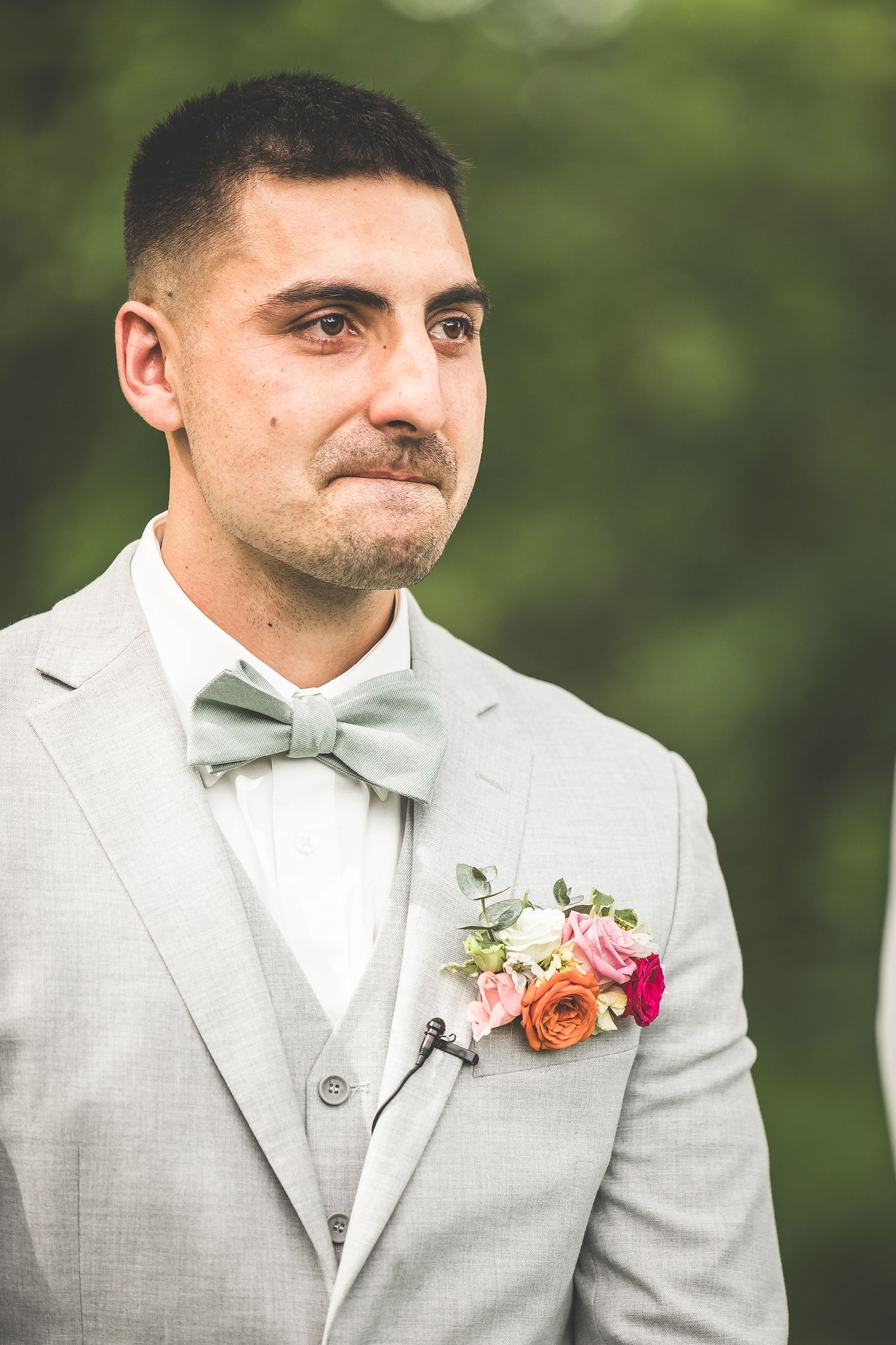 Groom in light gray suit with bow tie and floral boutonniere looking to the side outdoors.