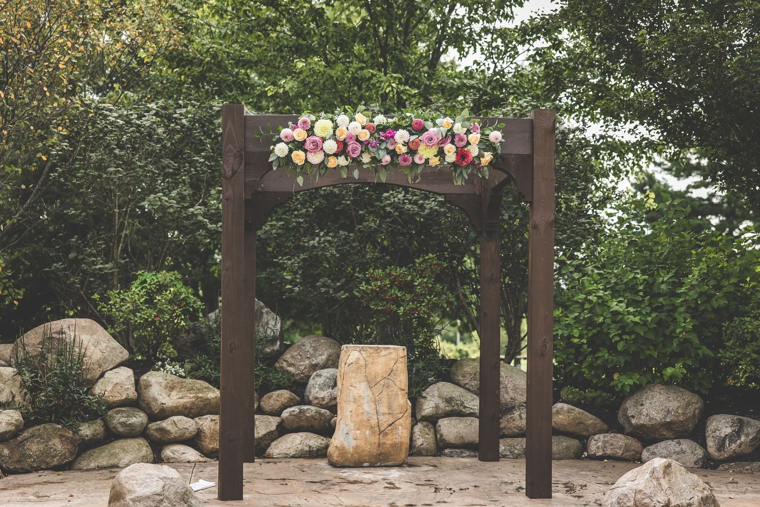 Wooden wedding ceremony arch decorated with pink and white flowers against stone wall.