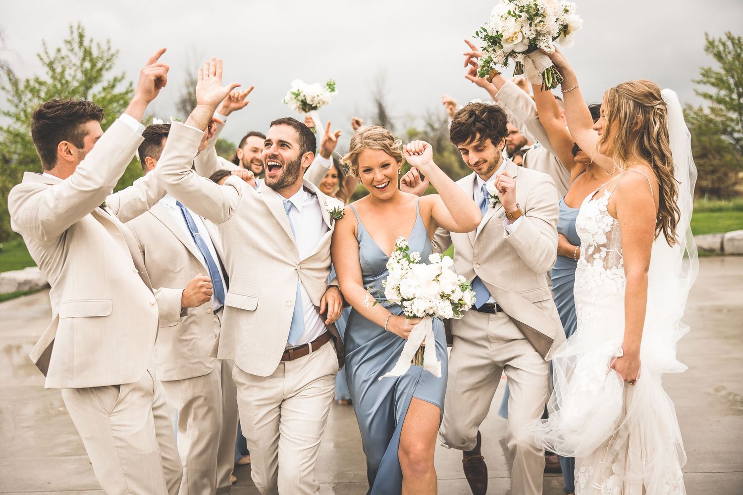 Wedding party celebrates joyfully outdoors wearing light beige suits and blue dresses with white bouquets.