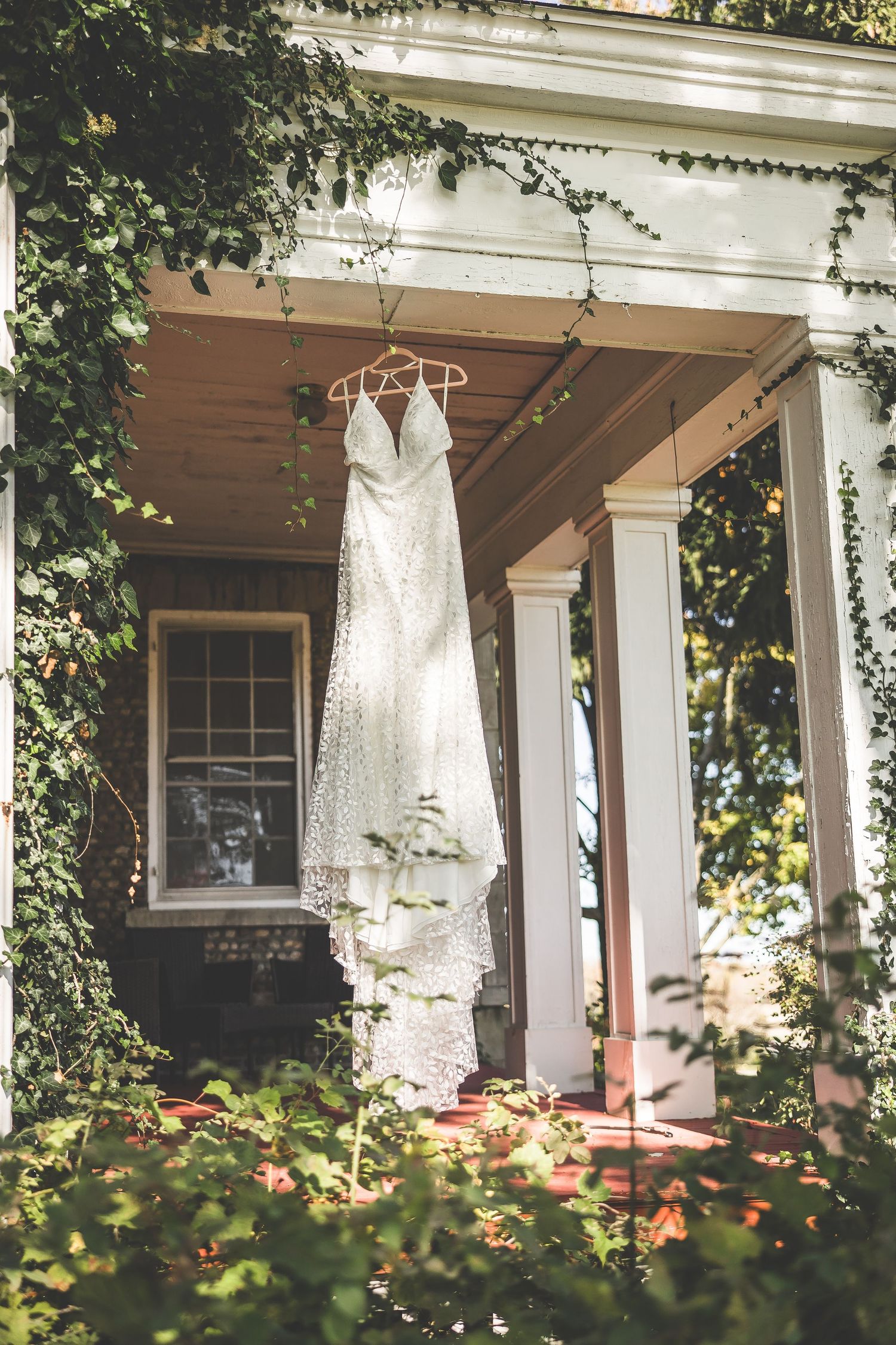 White wedding dress hanging on porch of classic white house with green foliage.