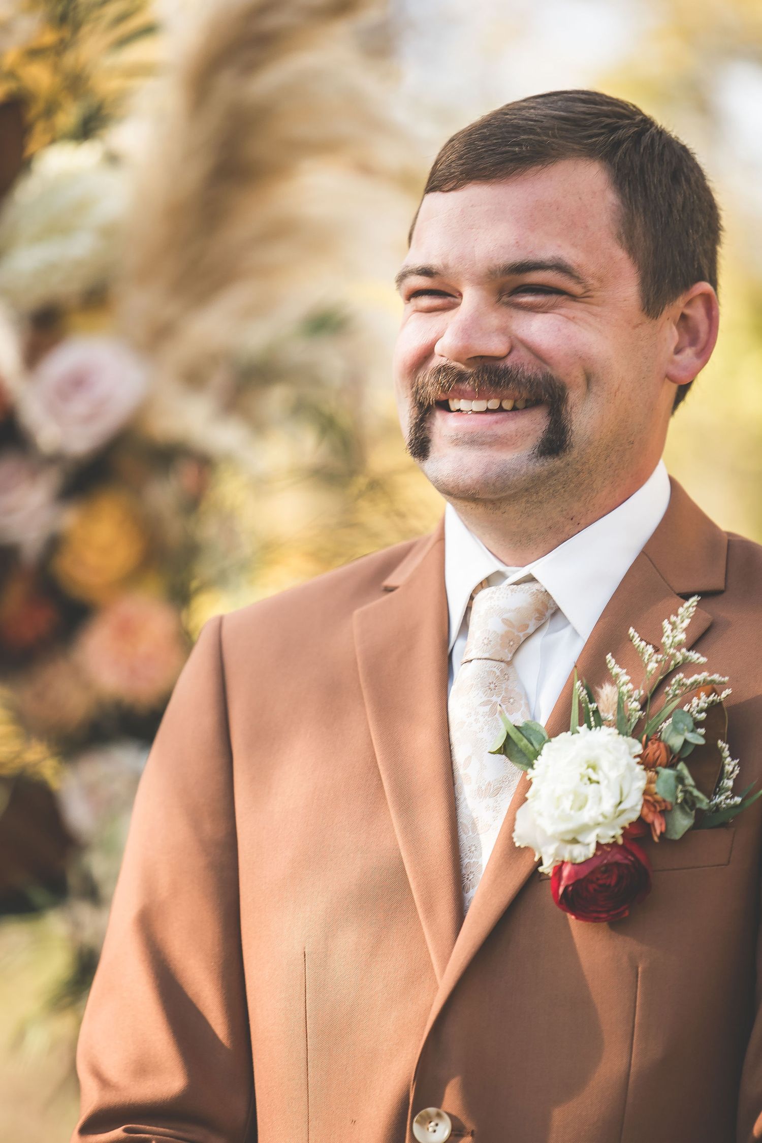 Groom in caramel-colored suit with white boutonniere smiles during outdoor autumn ceremony.