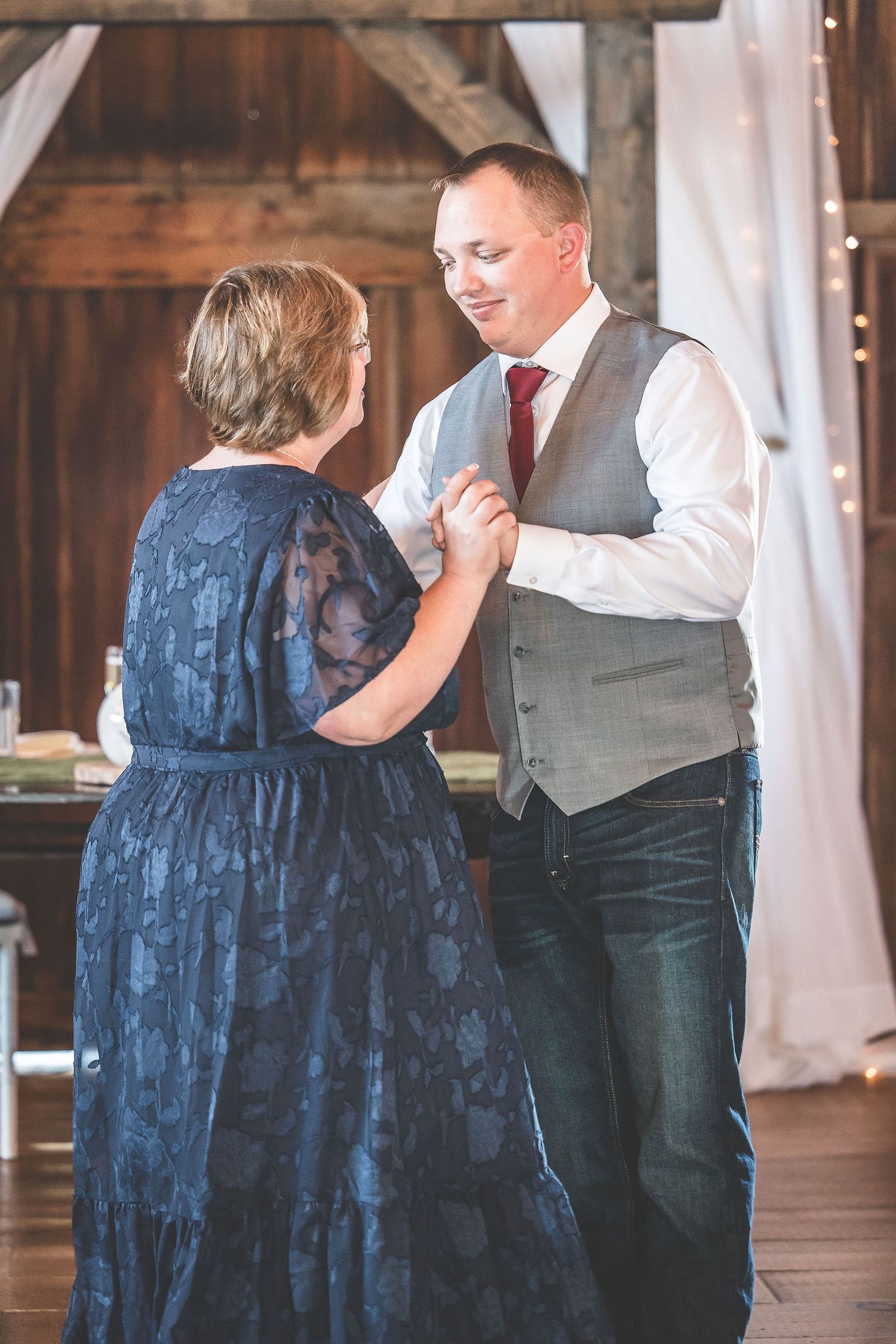 A couple shares an intimate dance moment in a rustic barn venue decorated with string lights.