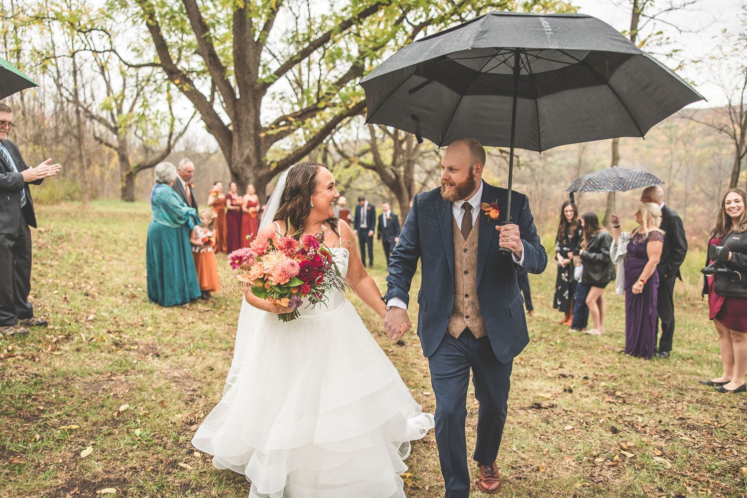 Wedding group gathers beneath large tree during autumn outdoor celebration.