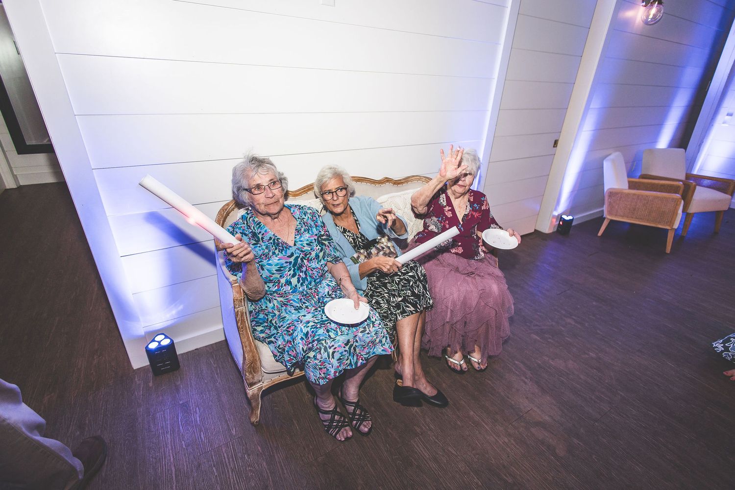 Elderly guests in colorful dresses wave light sticks while seated on a couch.