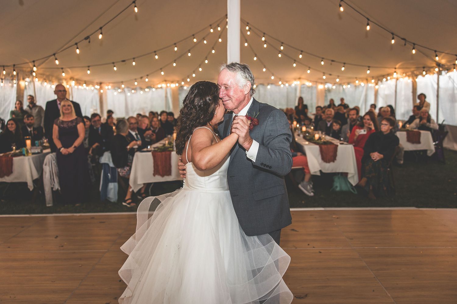 Couple performs wedding dance under string lights in white tent with wooden flooring.