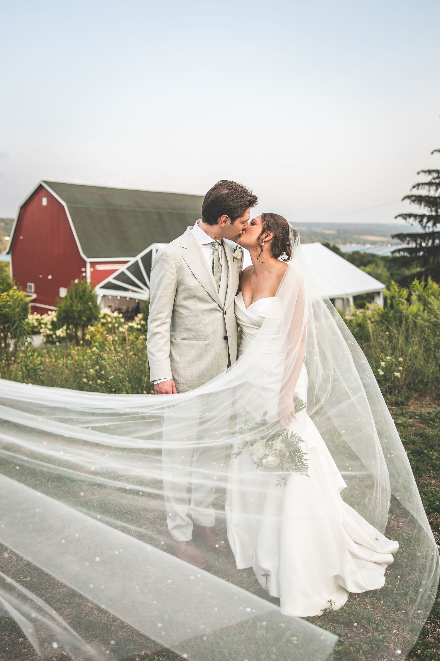 Flowing bridal veil catches the breeze during countryside wedding portraits near a red barn.