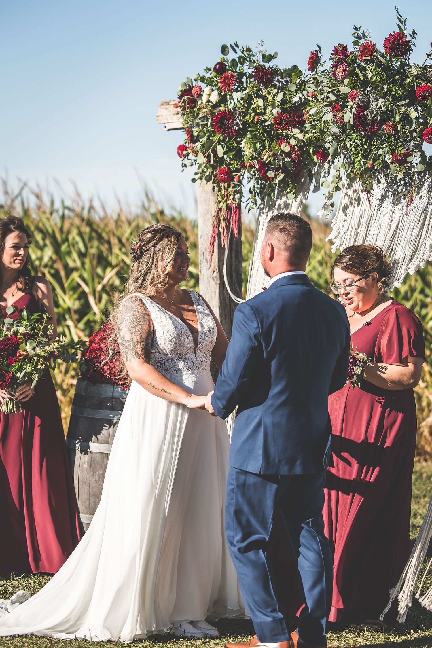 Wedding ceremony under floral arch with white macrame hangings and burgundy bridesmaids dresses.