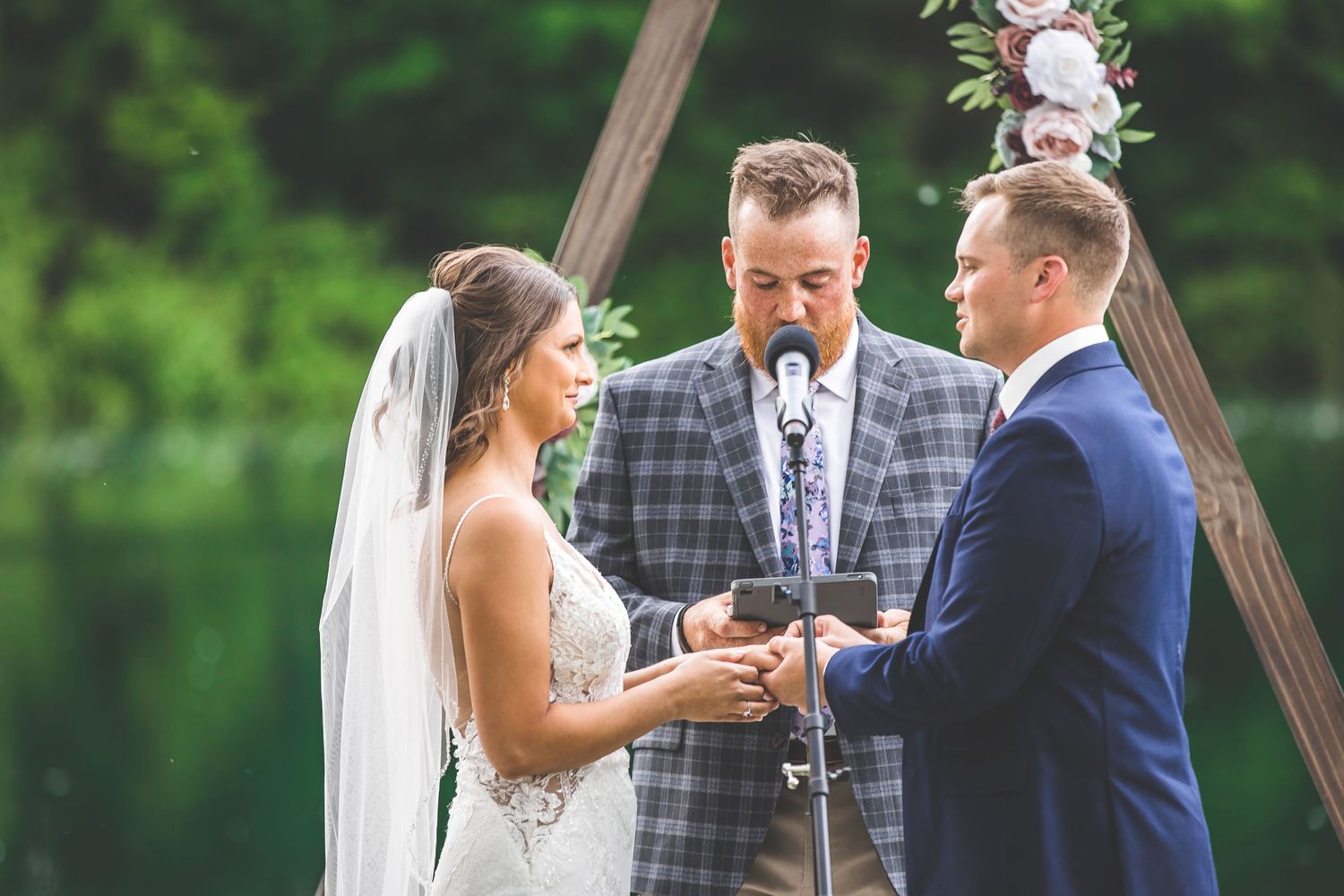 Outdoor wedding ceremony takes place under wooden arch with greenery in background.