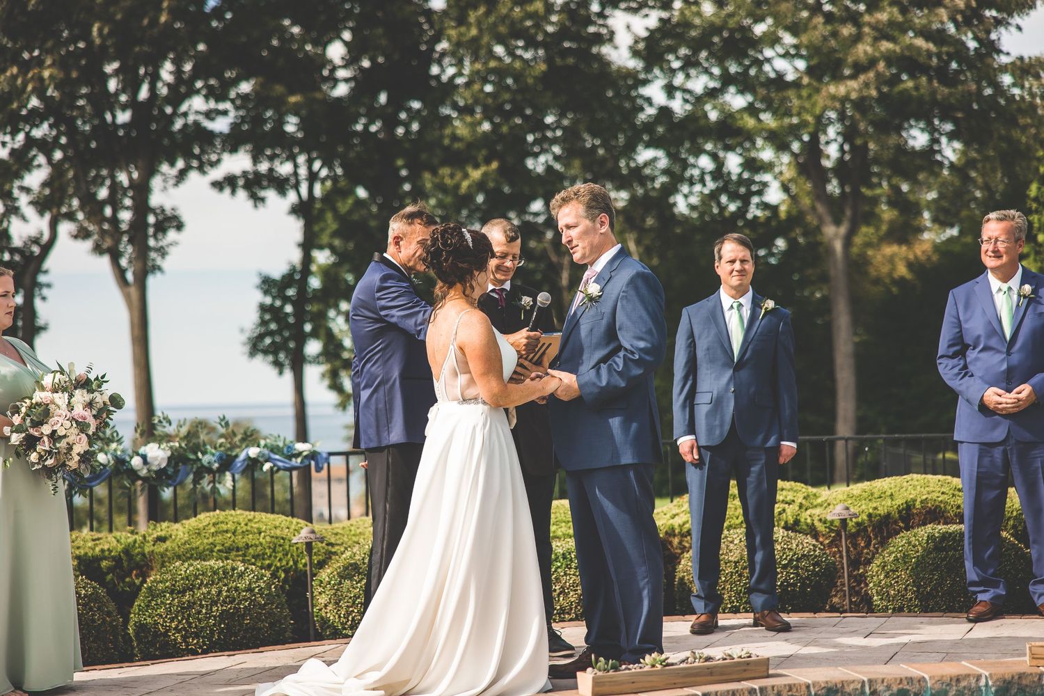 Wedding party gathered on stone terrace with manicured gardens and trees in background.