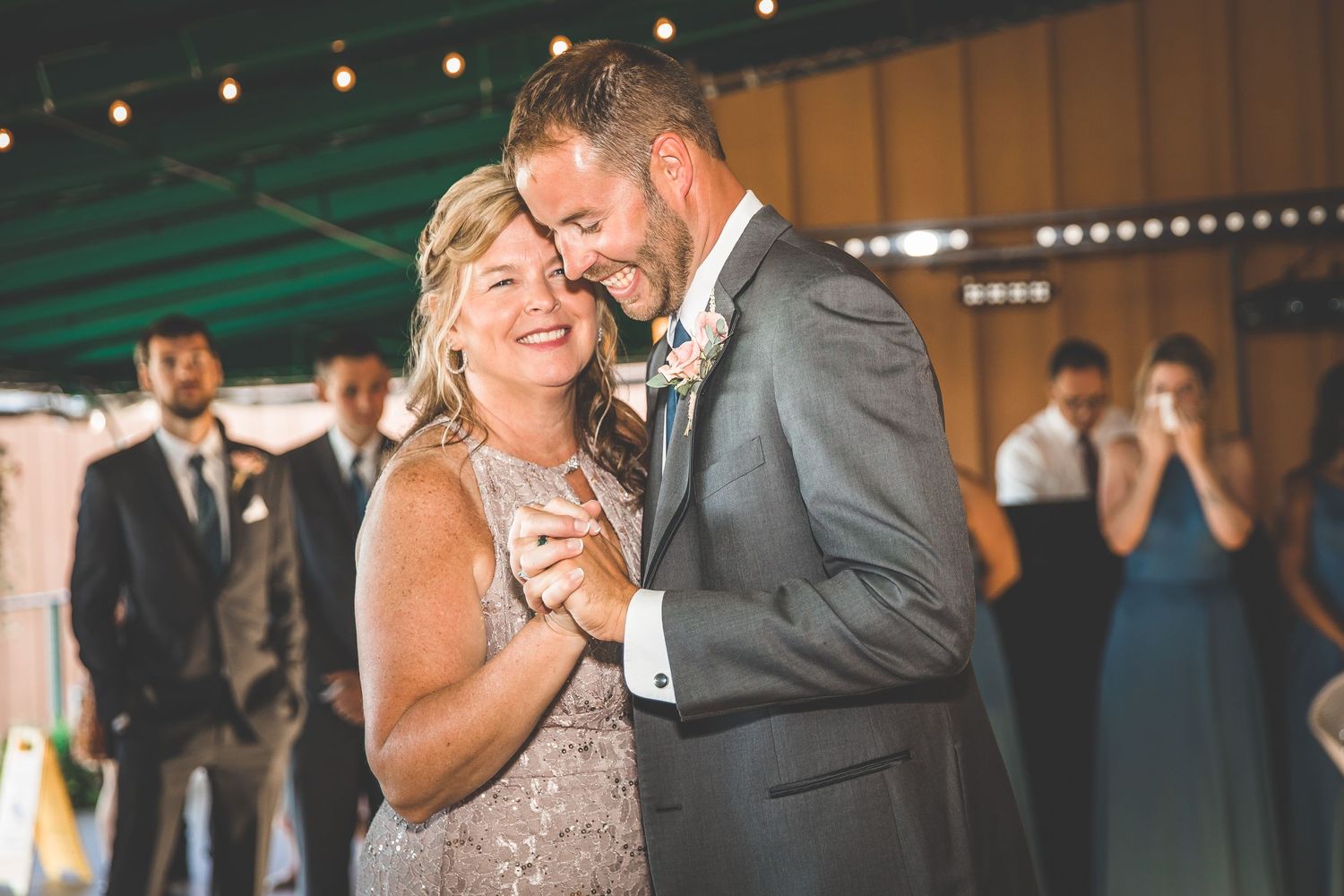 Wedding celebration under string lights with guests dancing in a rustic indoor venue.