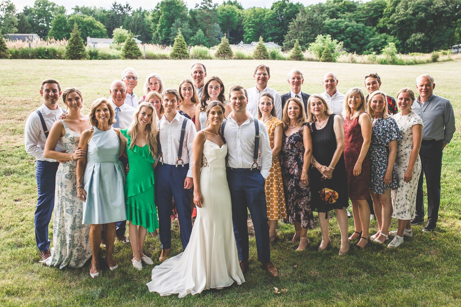 Large group photo of wedding guests posing together on a grassy lawn with trees in the background.