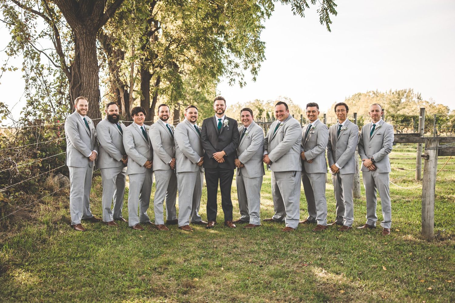 Groomsmen in light gray suits pose with groom in navy suit outdoors under autumn trees.