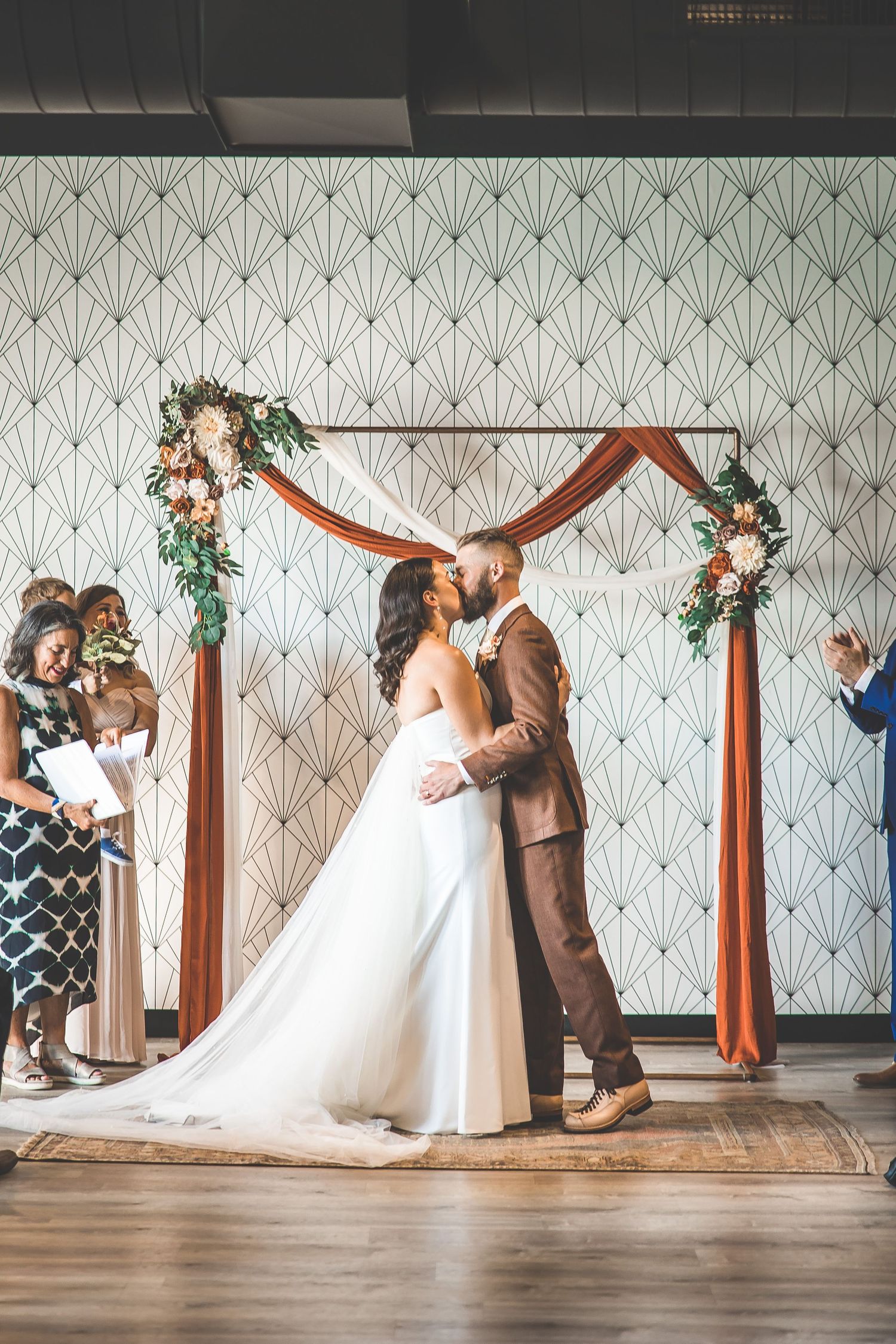 Wedding ceremony in modern venue with art deco wallpaper and wooden arch.