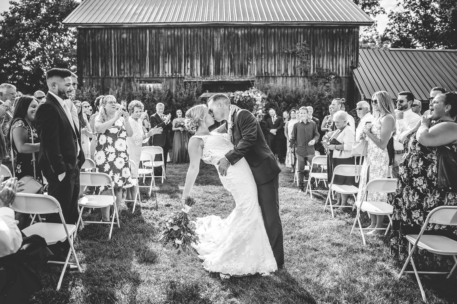 Wedding ceremony setup with white chairs on grass lawn in front of rustic barn building.