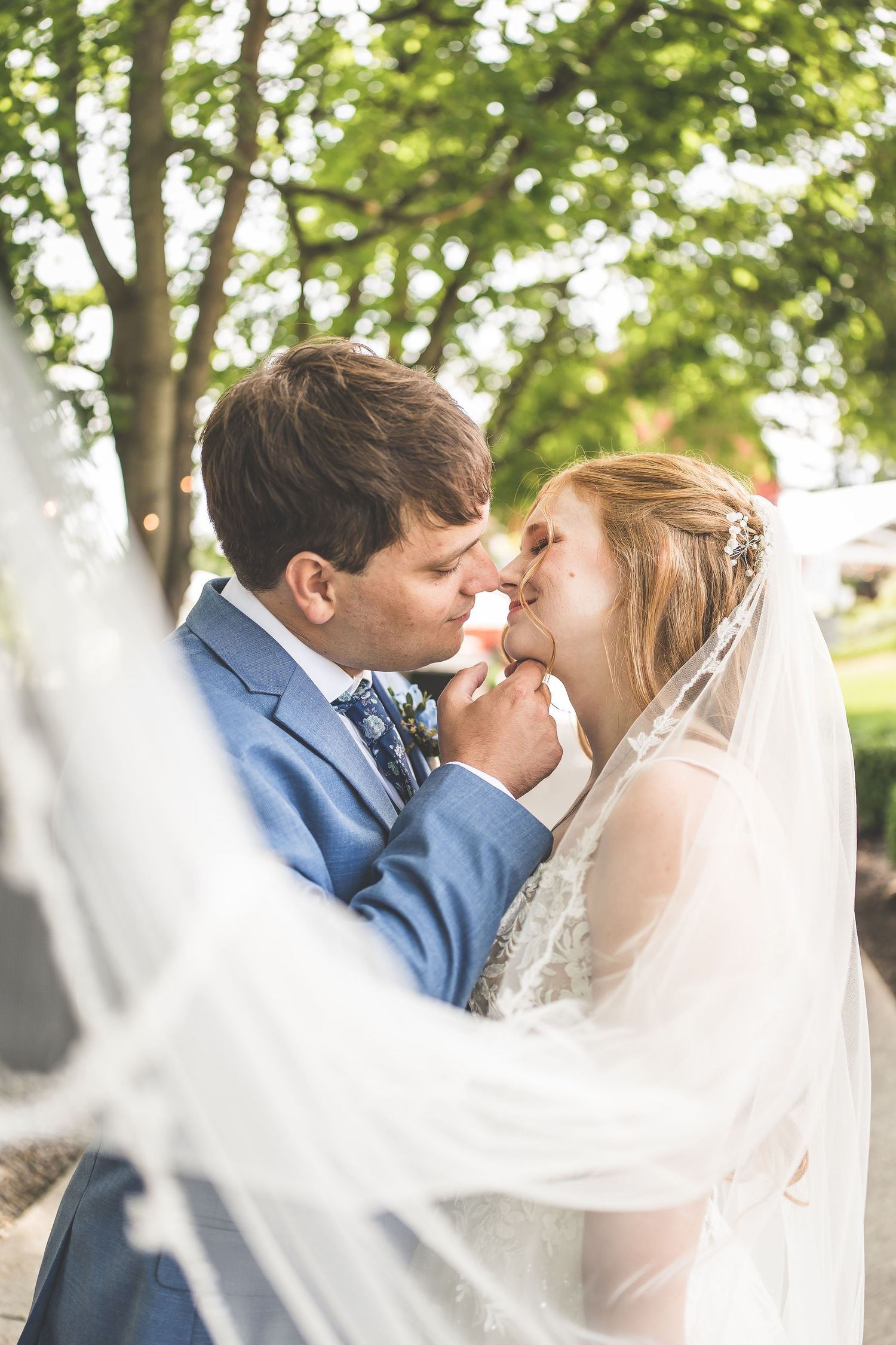 A bride and groom share a romantic moment under lush green trees, with the bride's veil flowing in the foreground.