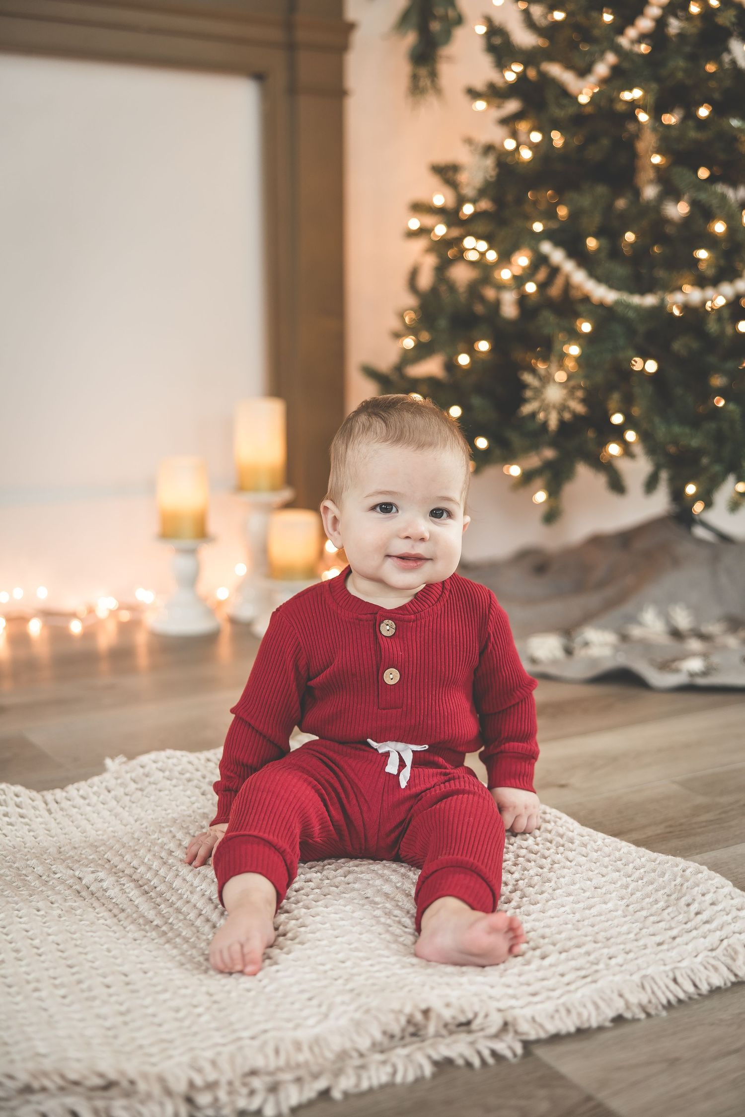 Baby wearing red knit outfit sitting on white blanket near Christmas tree.