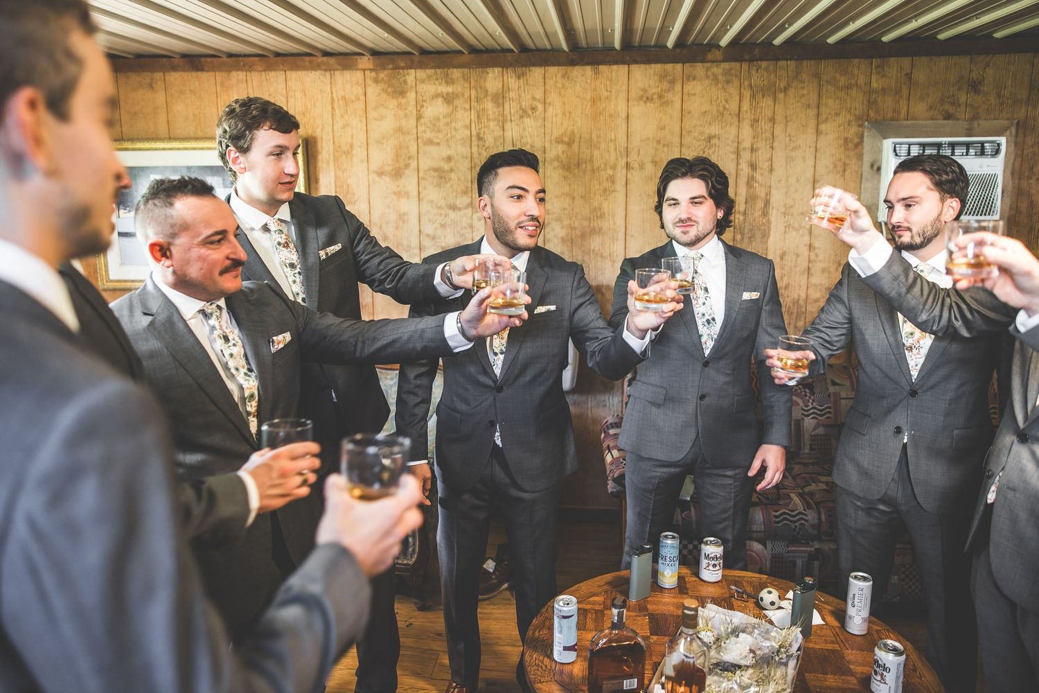 Groomsmen in dark suits raise glasses for celebratory toast in rustic indoor setting.