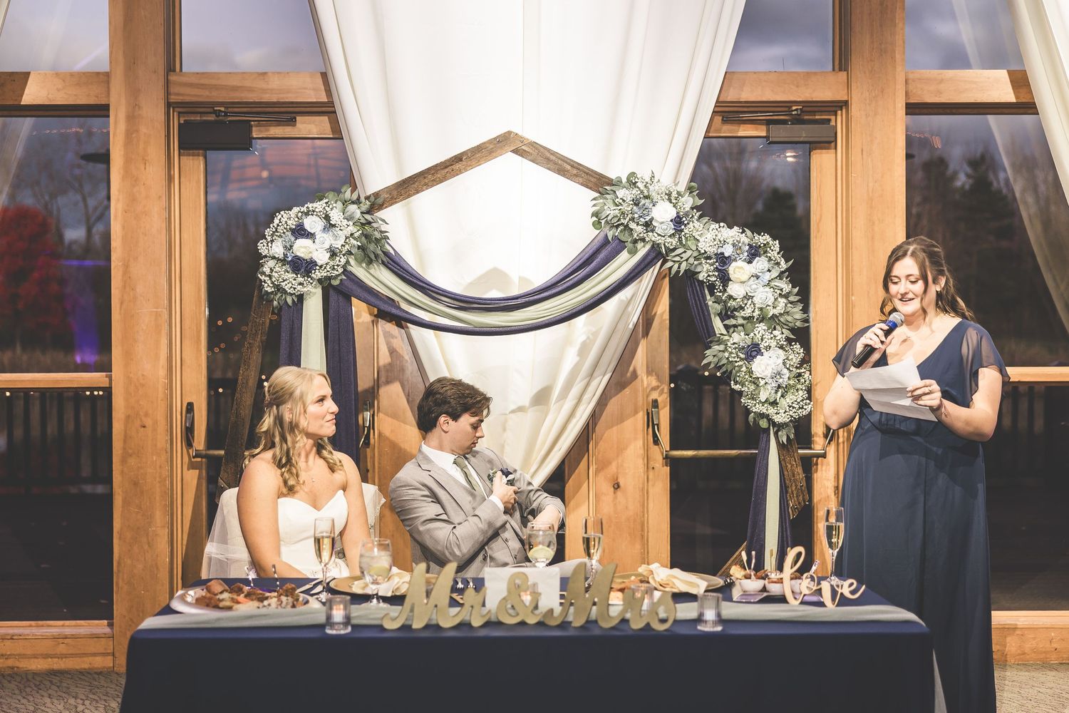 Wedding reception head table decorated with white floral arrangements and draped fabric.
