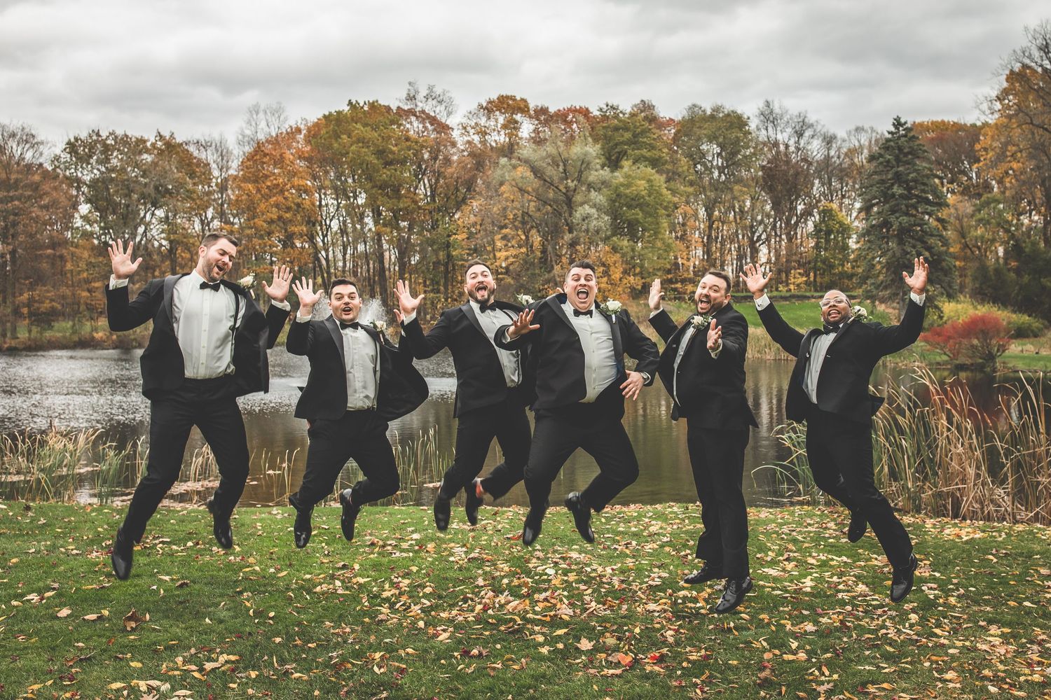 Groomsmen jumping and celebrating on lawn with fall trees in background.