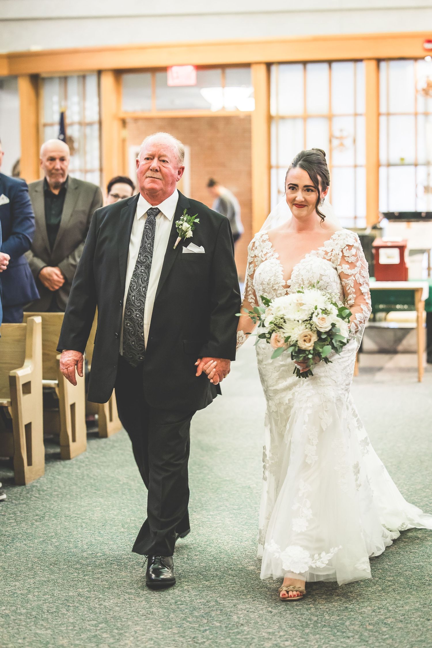 Wedding processional in church with companion escorting bride down carpeted aisle.