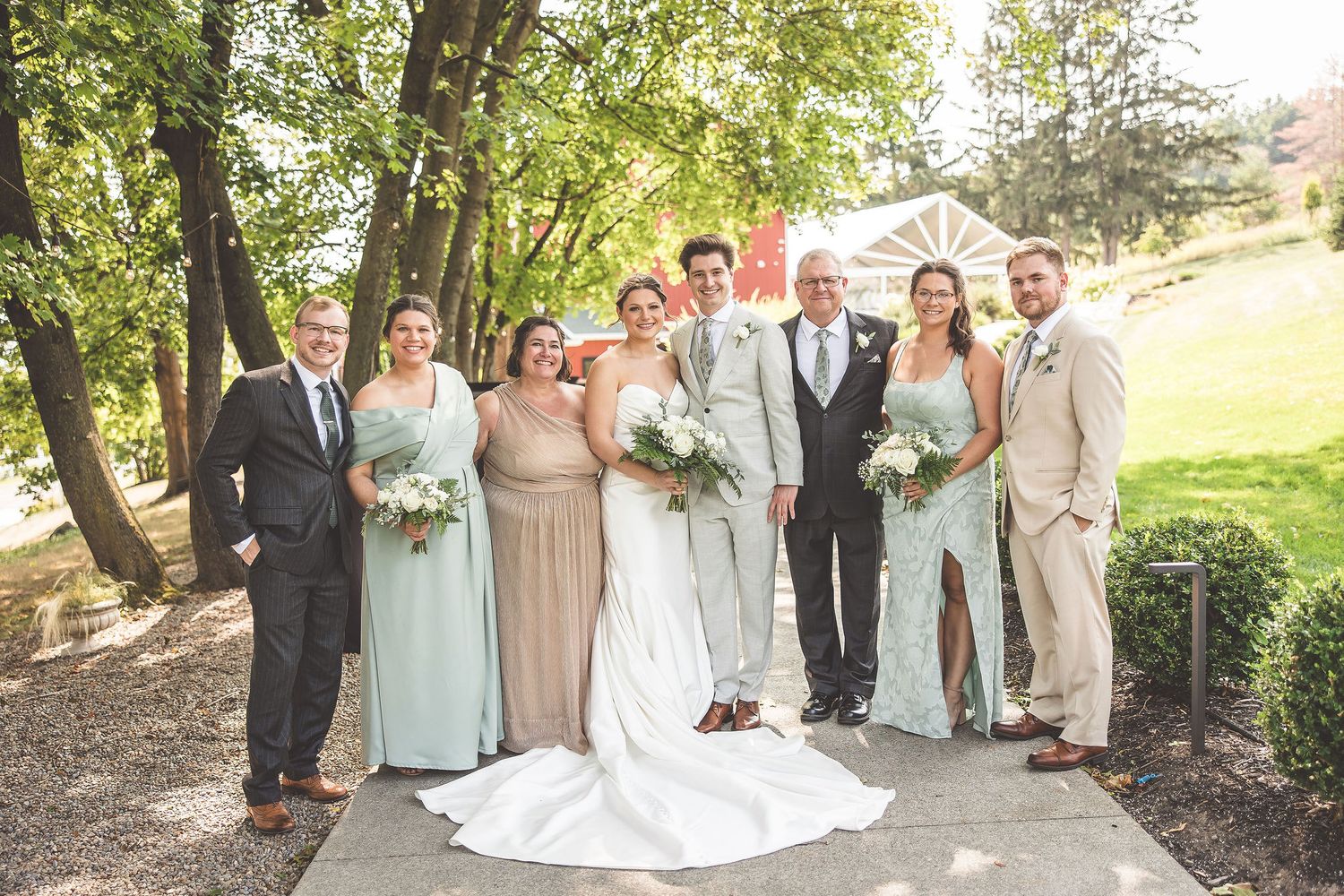 Wedding party poses outdoors on a tree-lined pathway for formal group photos.