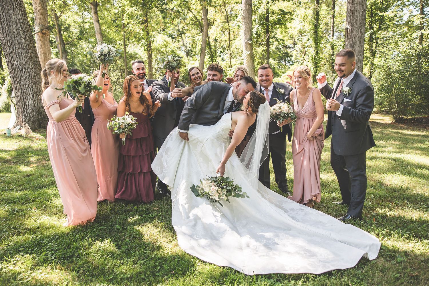 Wedding party celebrates with the couple's dramatic dress train displayed in a wooded setting.