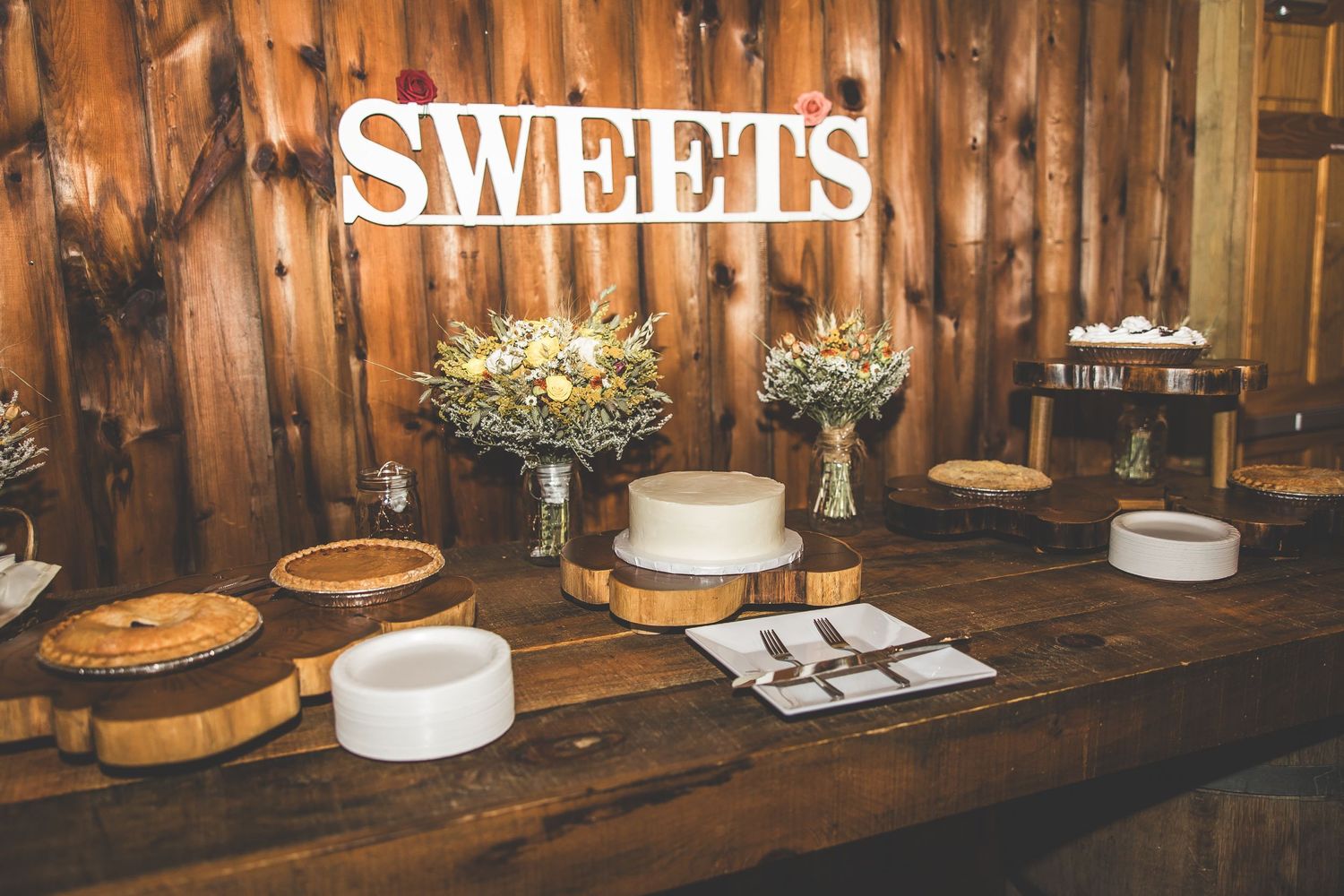 Dessert table with 'SWEETS' sign and multiple cakes displayed on wooden surface in rustic barn venue.
