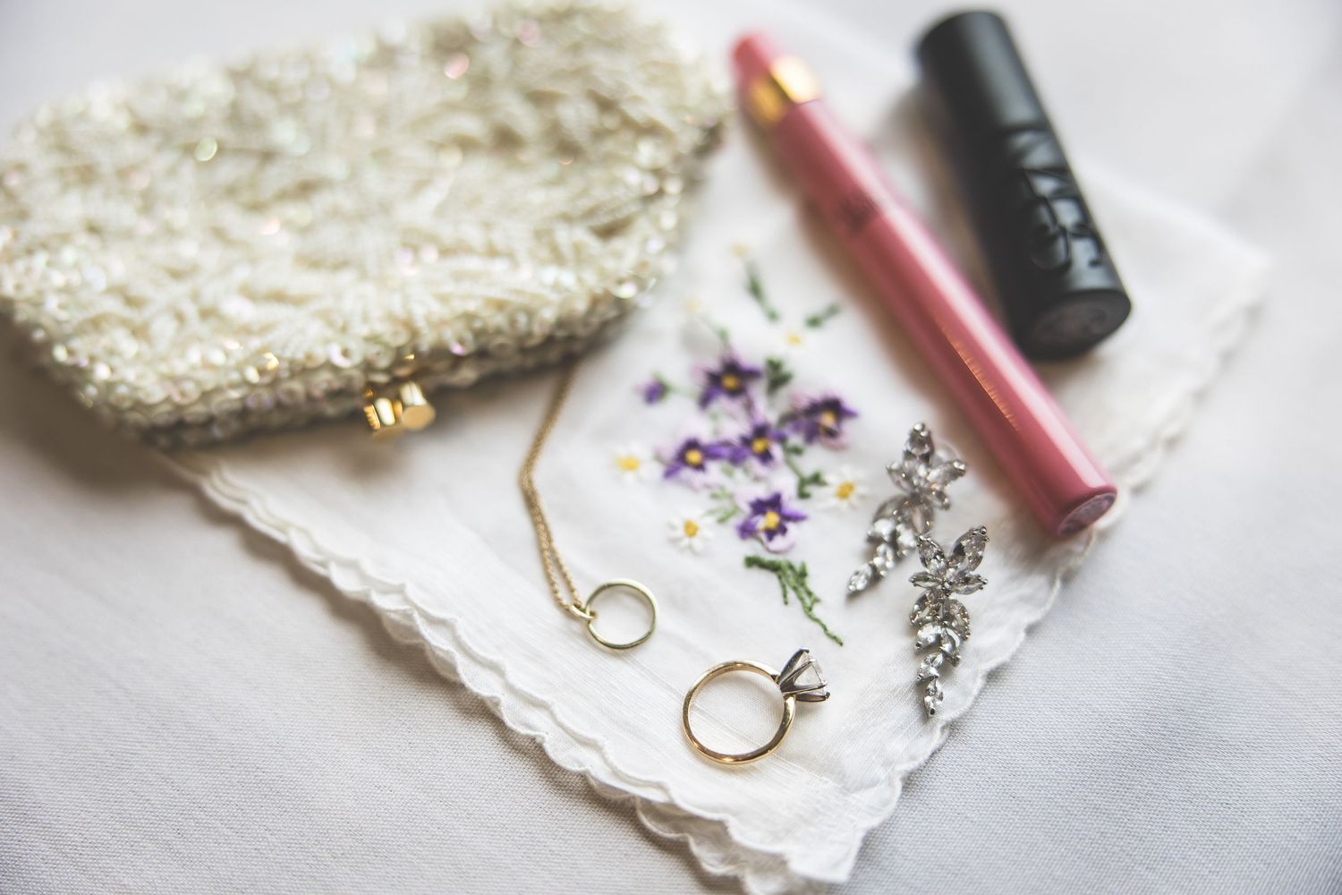 Wedding accessories including rings, lace handkerchief, and makeup items on white surface.