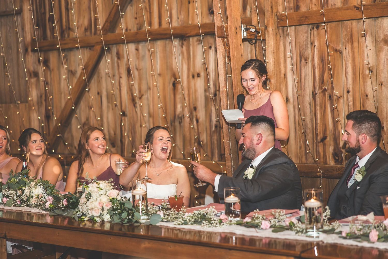 Long wooden tables set for wedding reception dinner in rustic barn venue with string lights.