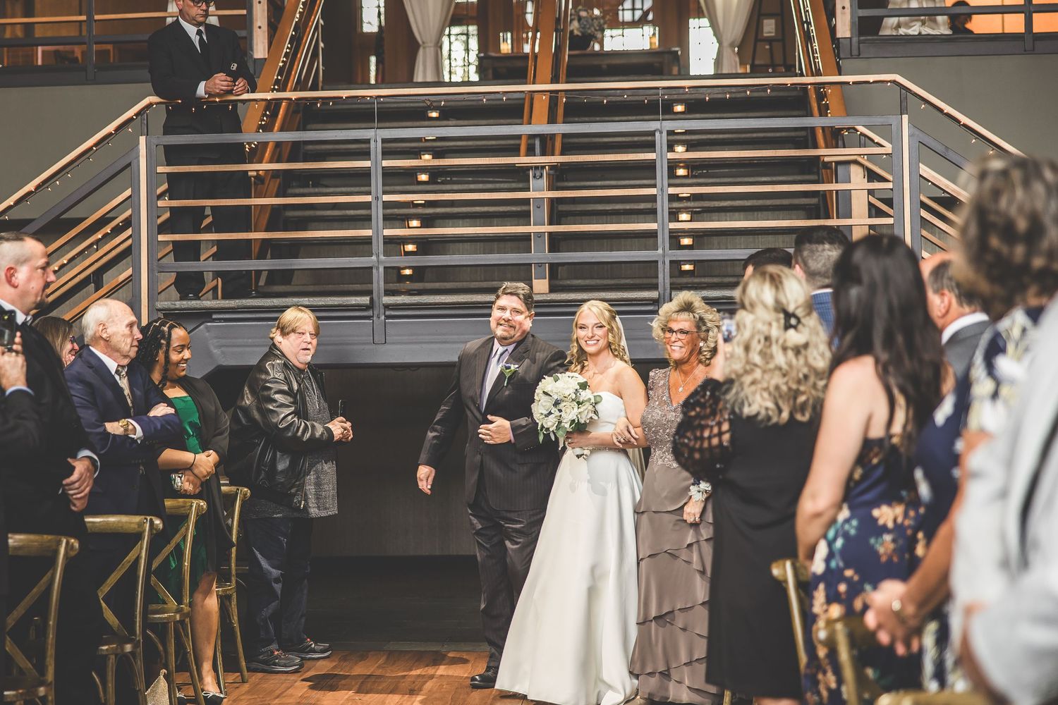 Wedding ceremony guests gather in an industrial-style venue with wooden architecture and white drapery.
