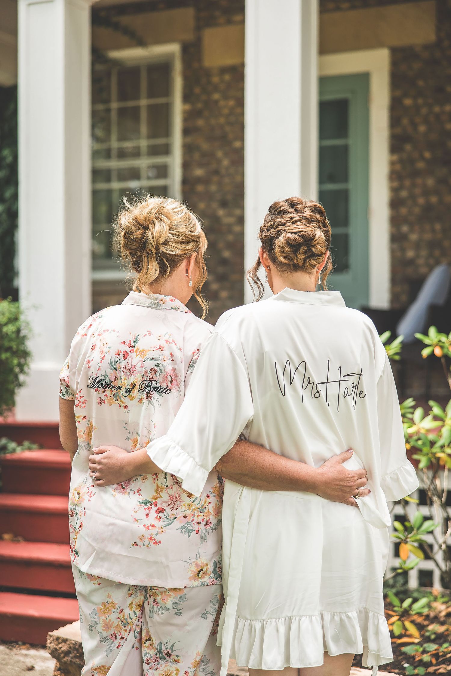 Two people wearing floral and white robes embracing from behind on porch steps.