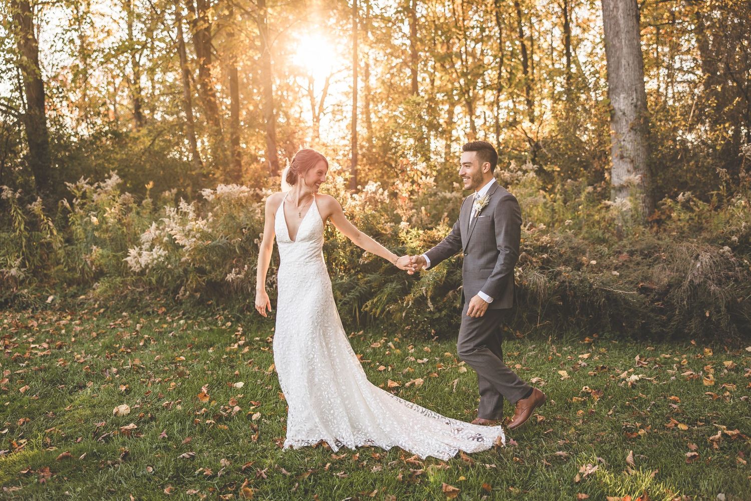 Couple holding hands in autumn forest with sunlight streaming through trees during wedding photoshoot.