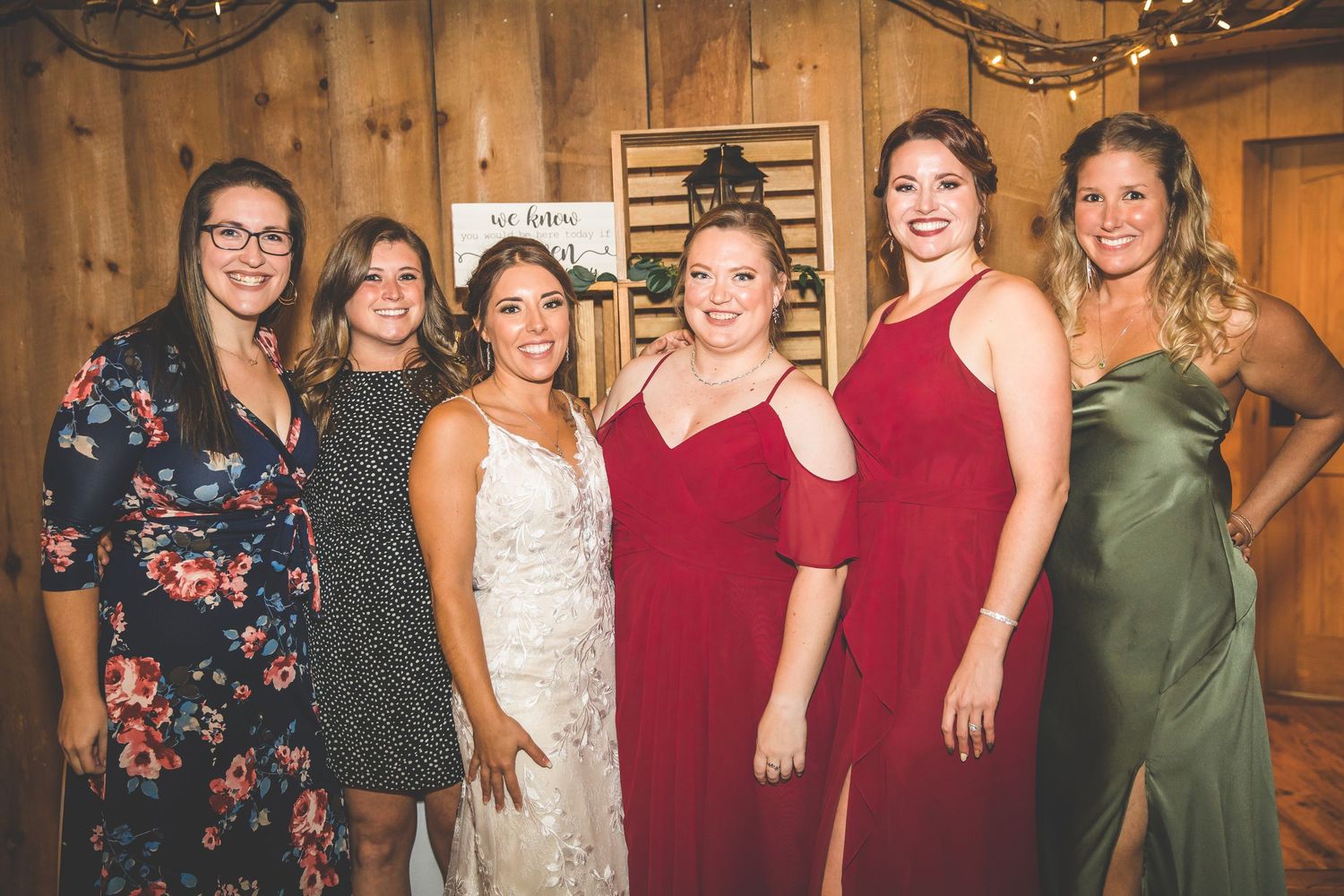 Wedding party poses together in rustic barn venue with string lights and wooden backdrop.