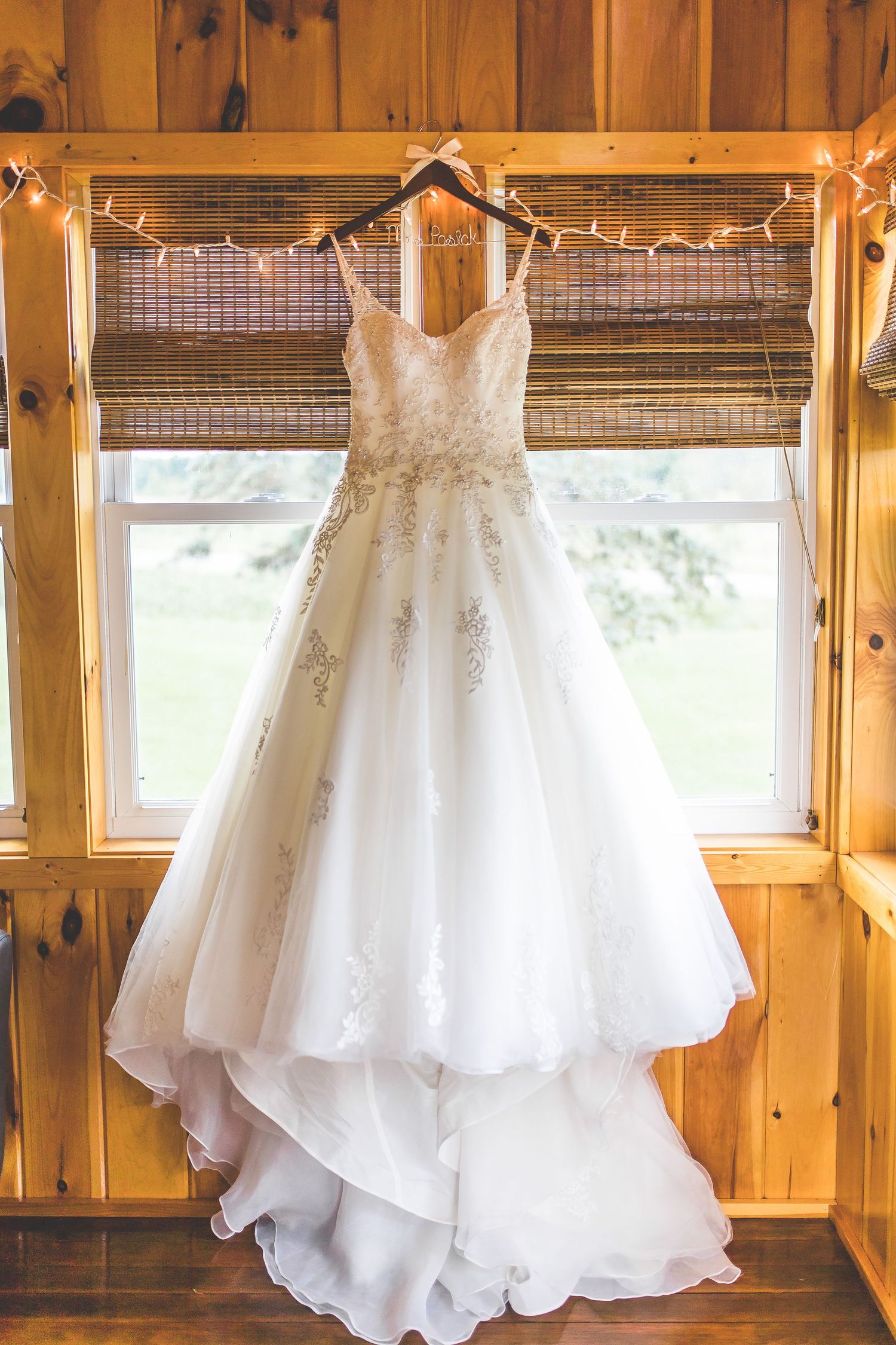 A white lace wedding dress hangs in a rustic wooden window frame decorated with string lights.