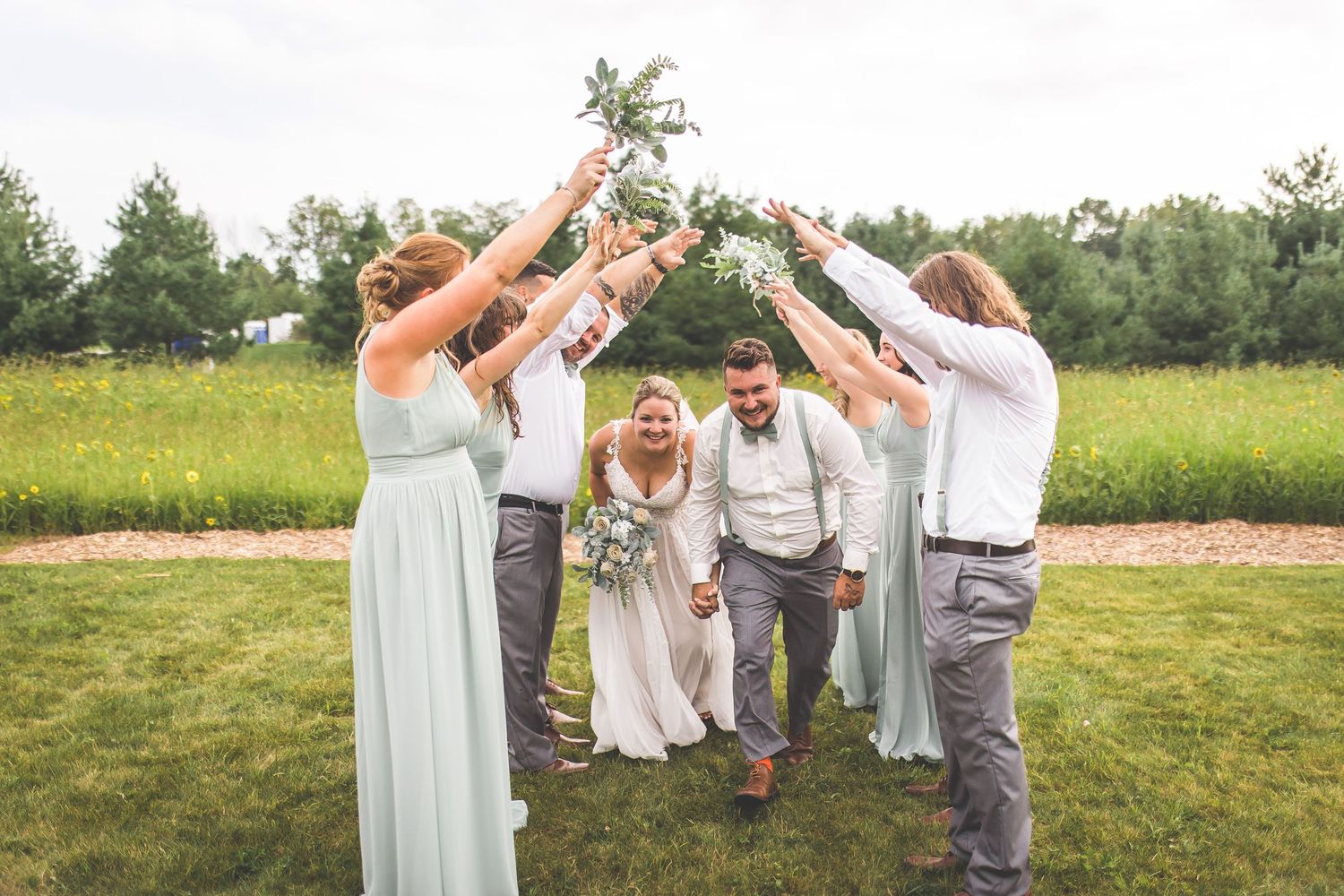 Wedding party poses playfully outdoors with groomsmen in suspenders and bridesmaids in sage dresses.