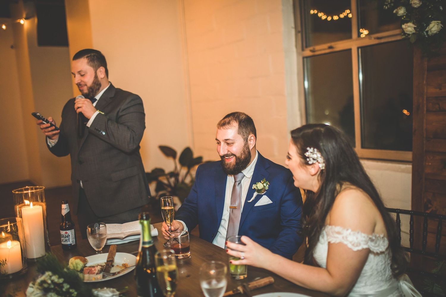 Wedding reception scene with guests enjoying dinner and drinks under warm lighting and string lights.