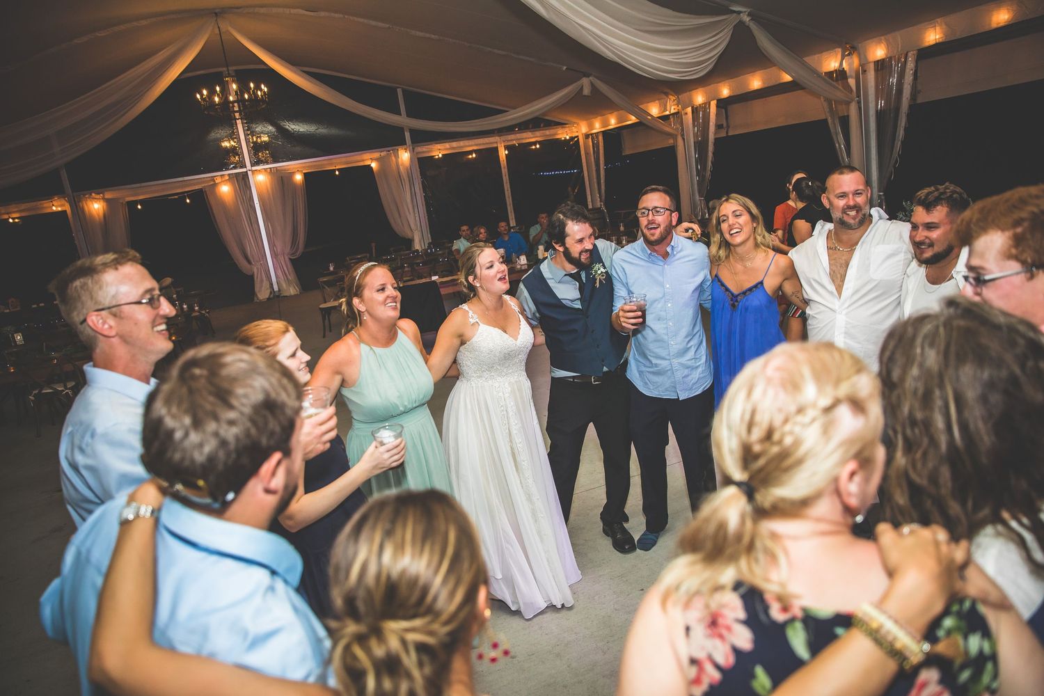Wedding guests dancing and celebrating under a tent decorated with white draping and lights.