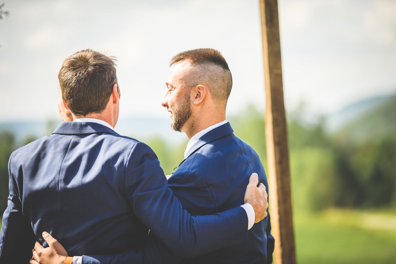 Wedding guests gather for an outdoor ceremony under wooden posts with floral decorations.