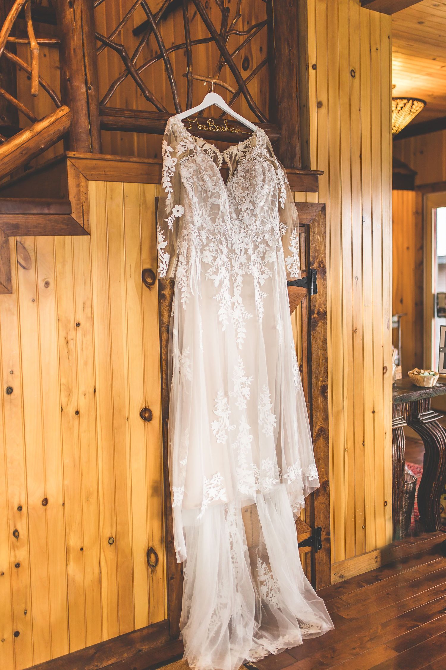 An elegant lace wedding dress with long sleeves hangs on display against wooden cabin walls.
