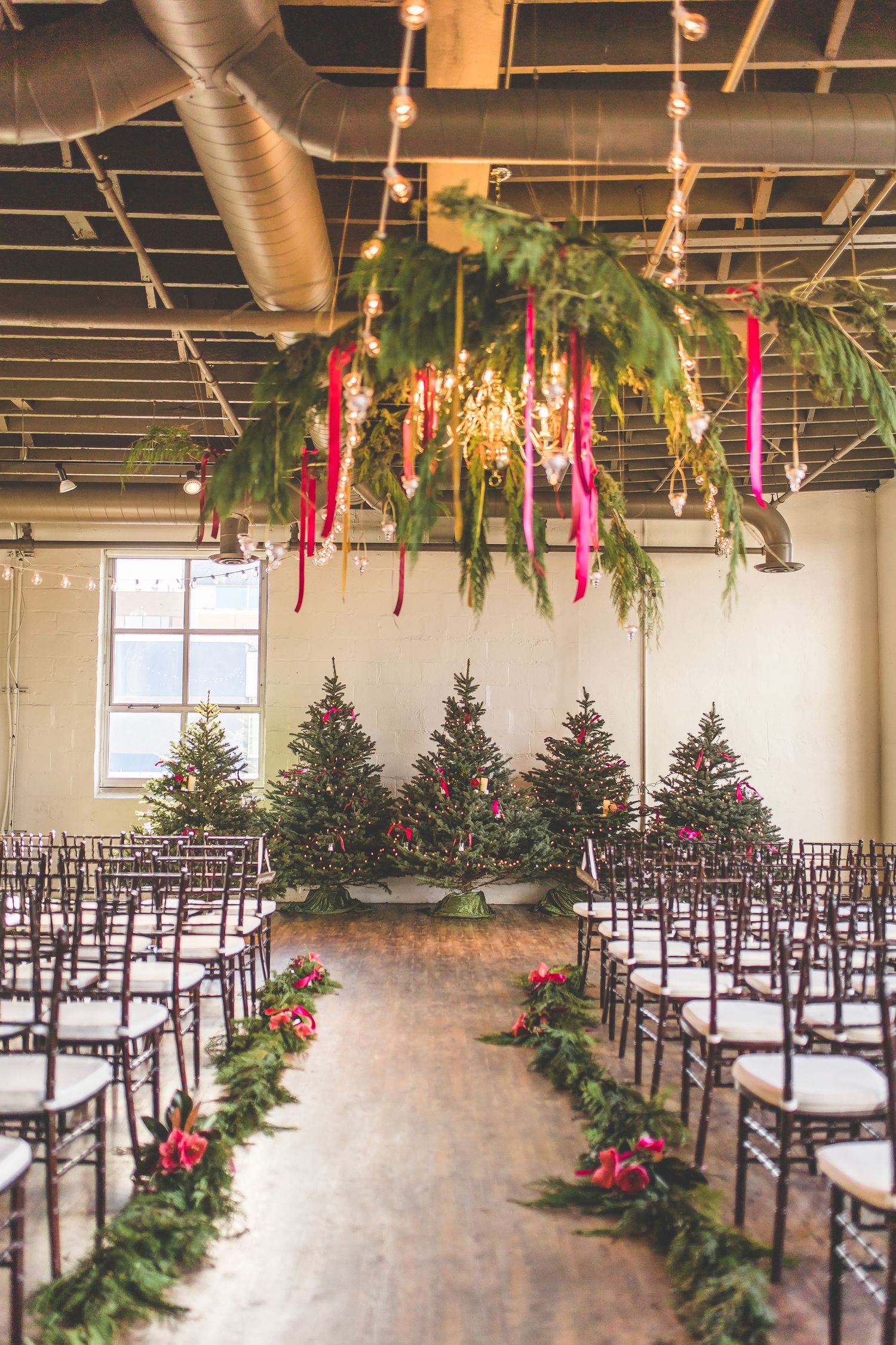 Indoor wedding ceremony setup with Christmas trees and hanging colorful ribbons in an industrial space.
