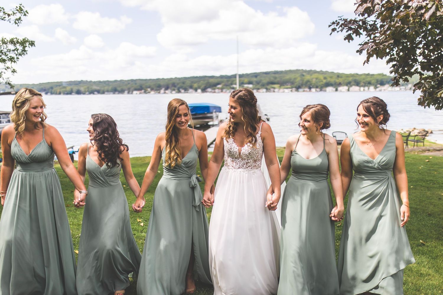 Wedding party in sage green dresses poses together on grassy lawn by waterfront.