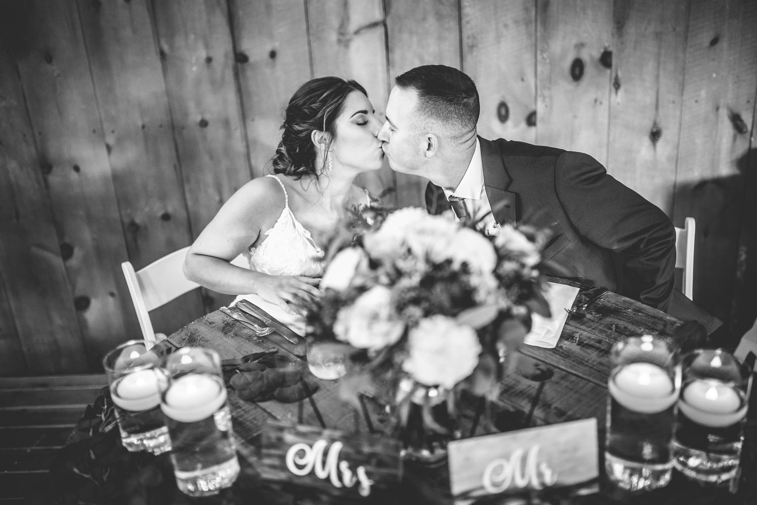 Black and white photo of a couple sharing an intimate moment at their reception table with candles and flowers.