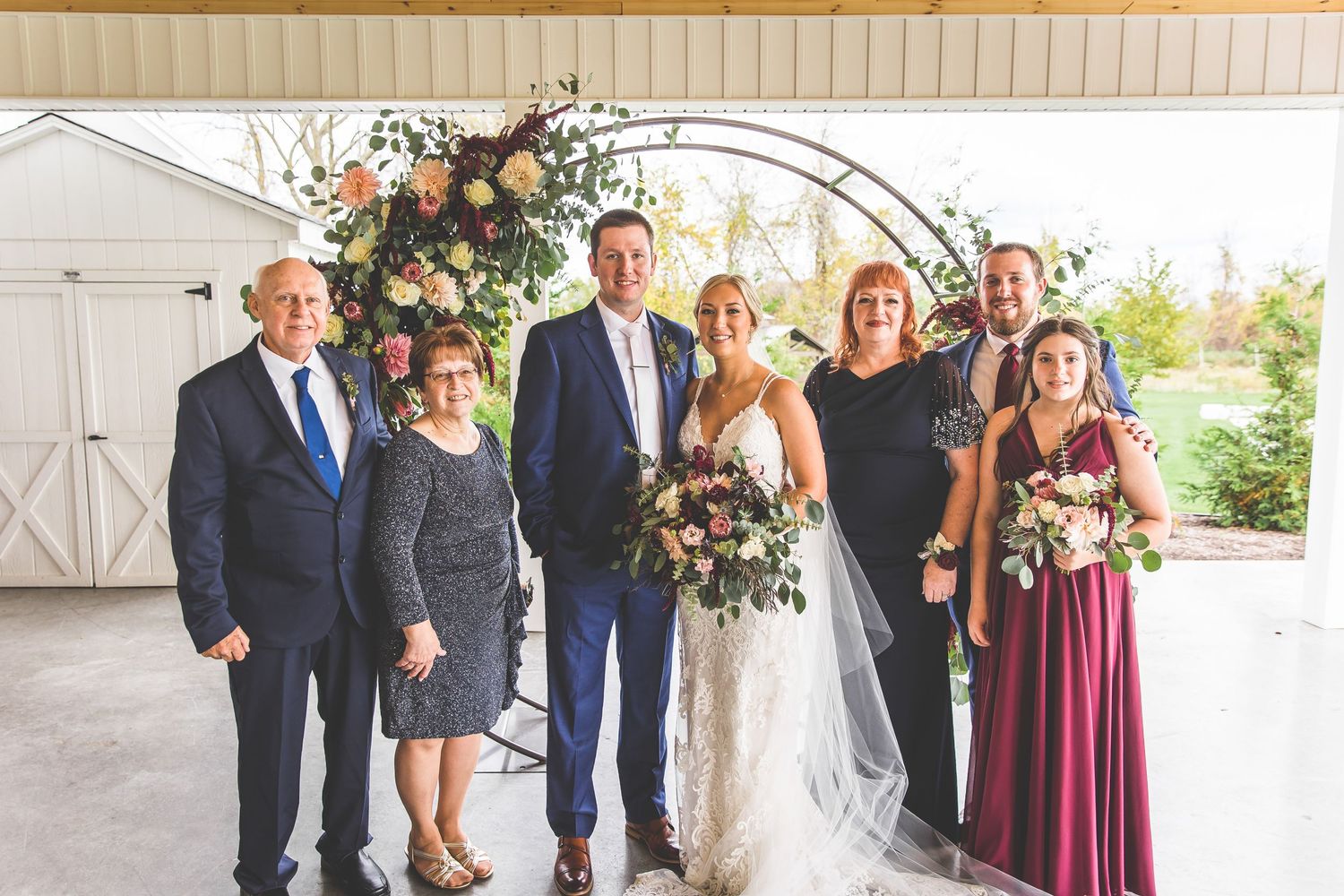 Wedding party poses under floral arch with burgundy and navy color scheme.
