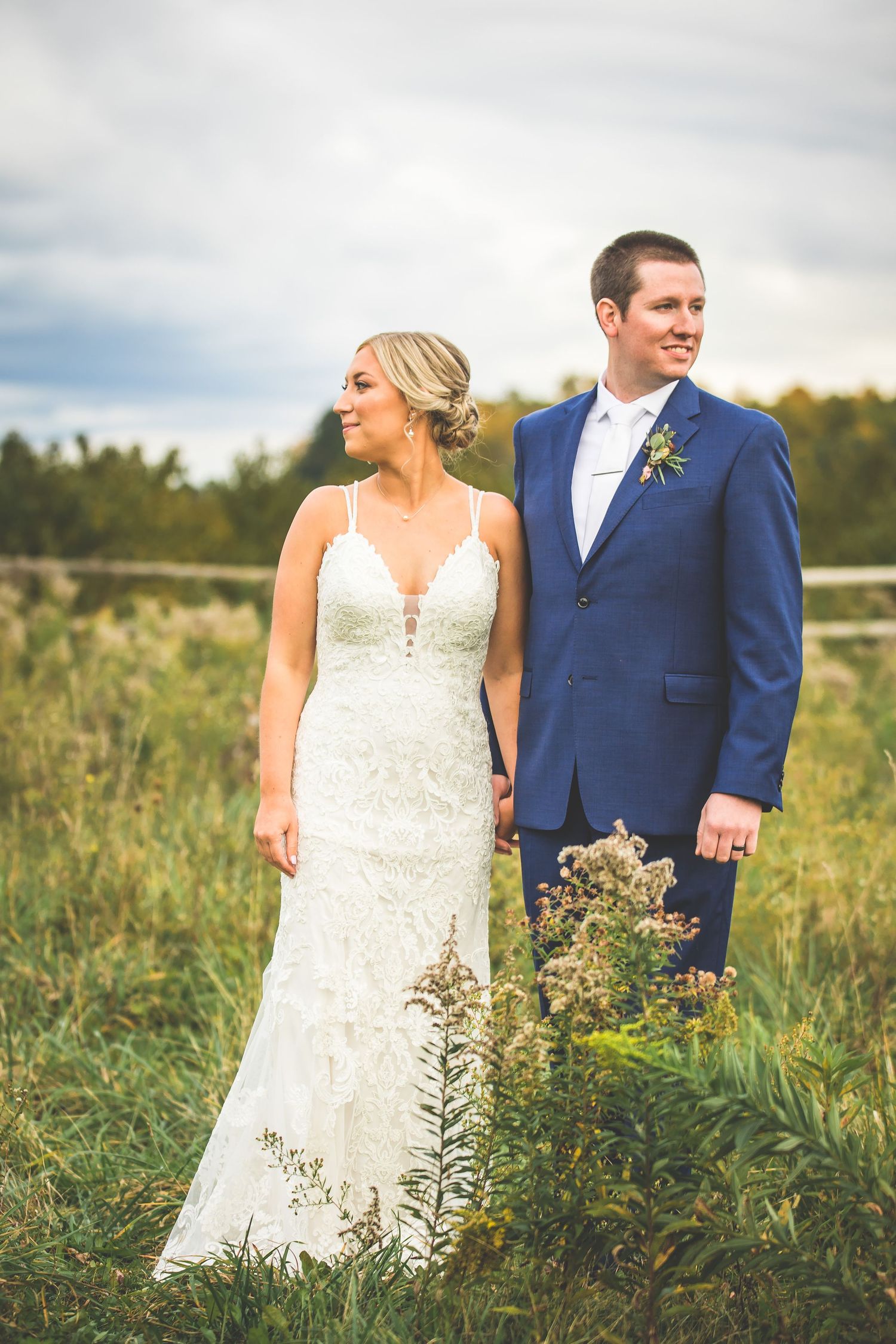 Wedding couple stands in tall grass field wearing blue suit and white lace dress.