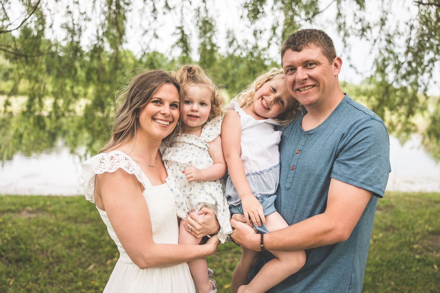 Happy family portrait outdoors with lake and willow trees in background.