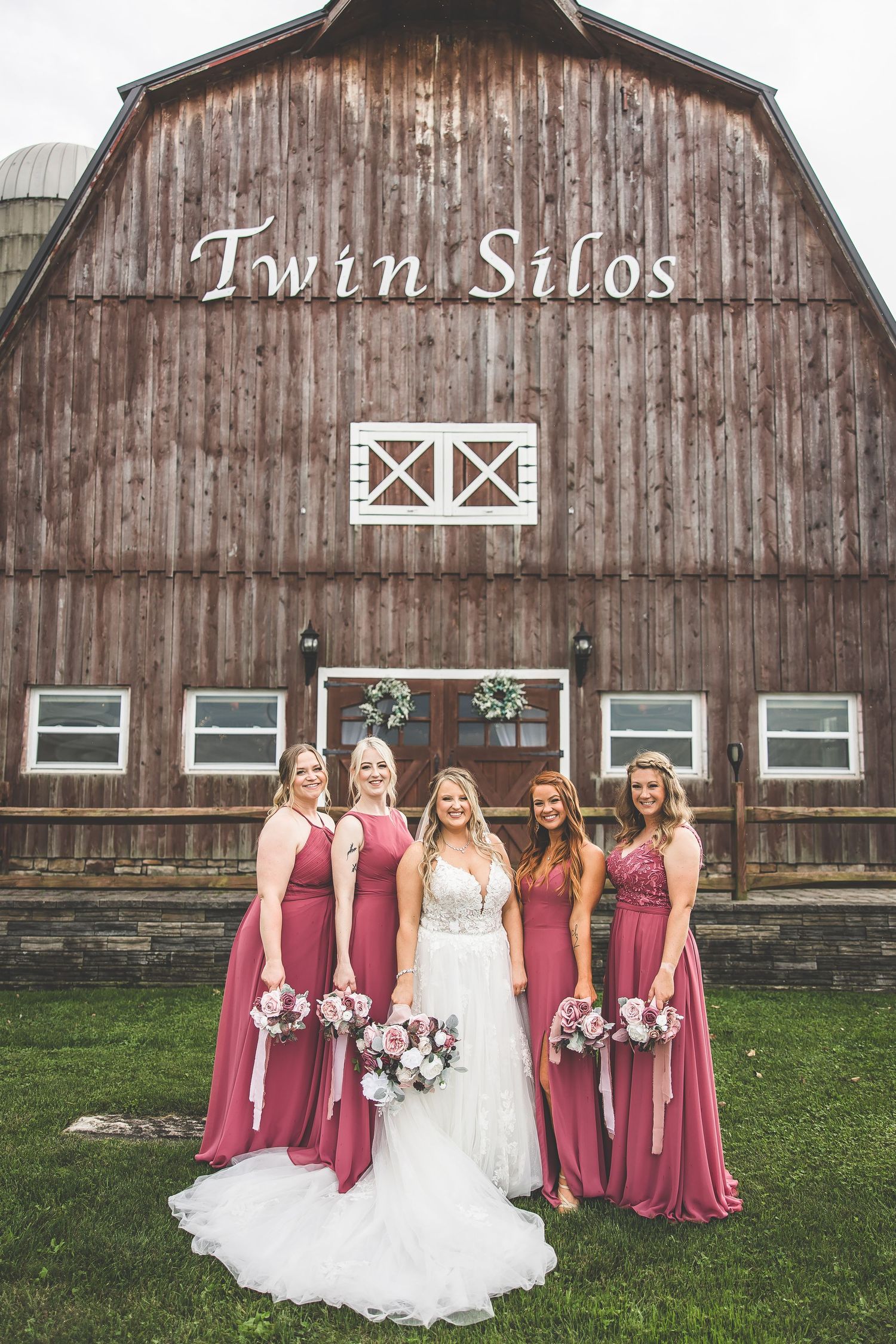 Wedding party poses in front of rustic Twin Silos barn with bridesmaids in dusty rose dresses.