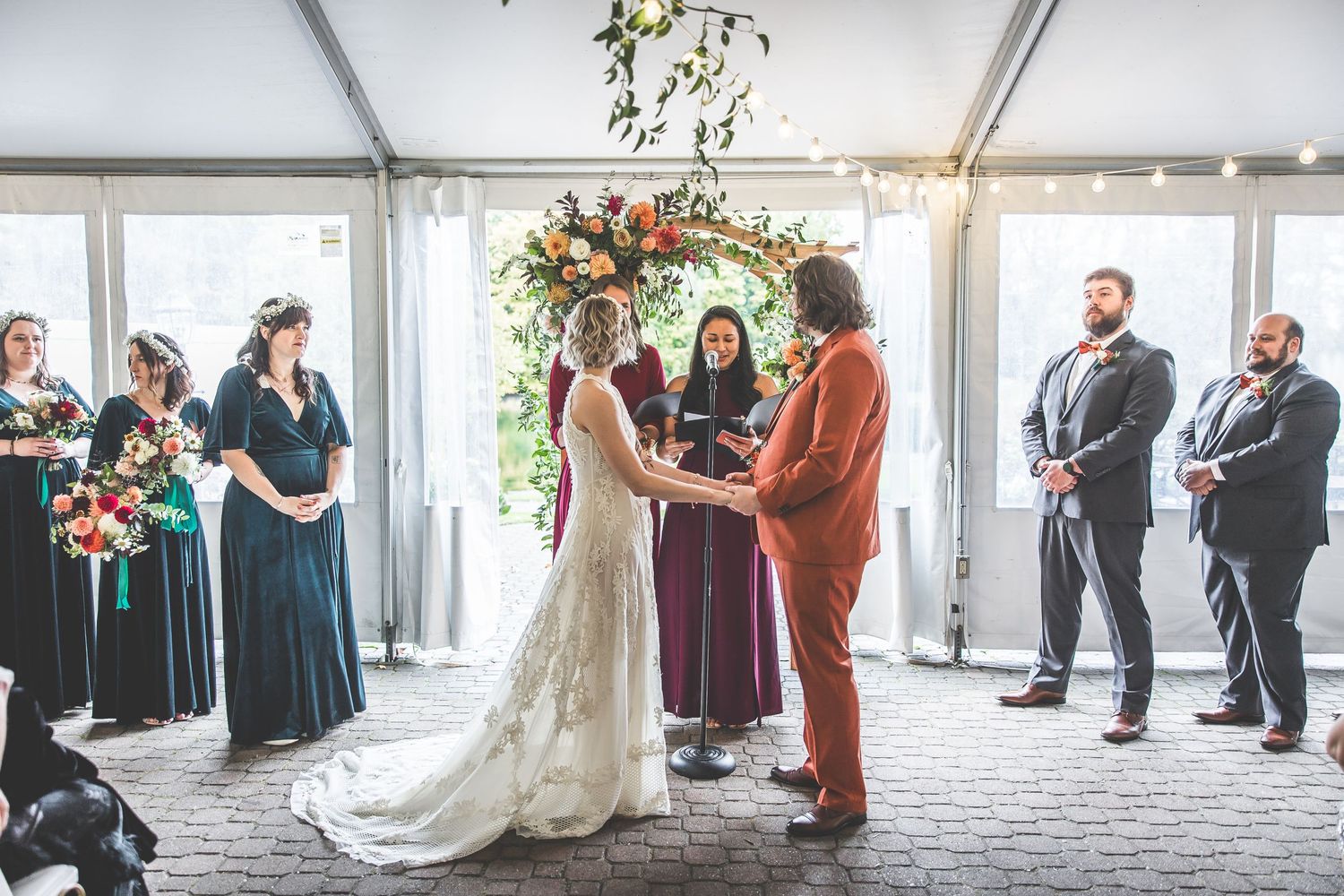 Indoor wedding ceremony with guests seated in rows under string lights and greenery.