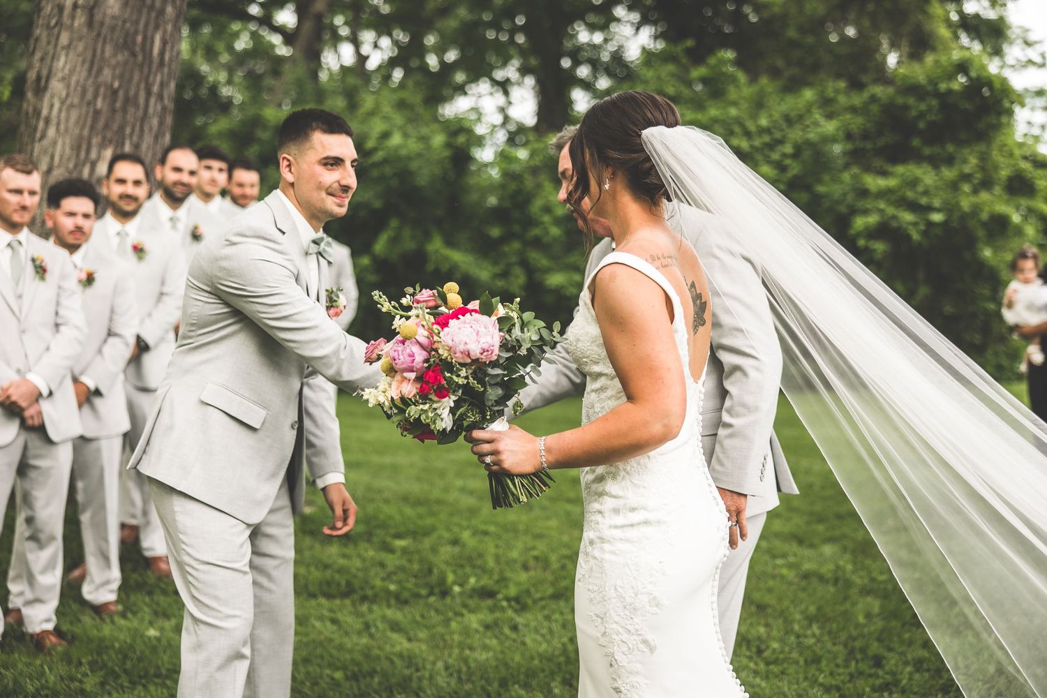 Joyful wedding couple exchanging bouquet during outdoor ceremony with groomsmen in background.
